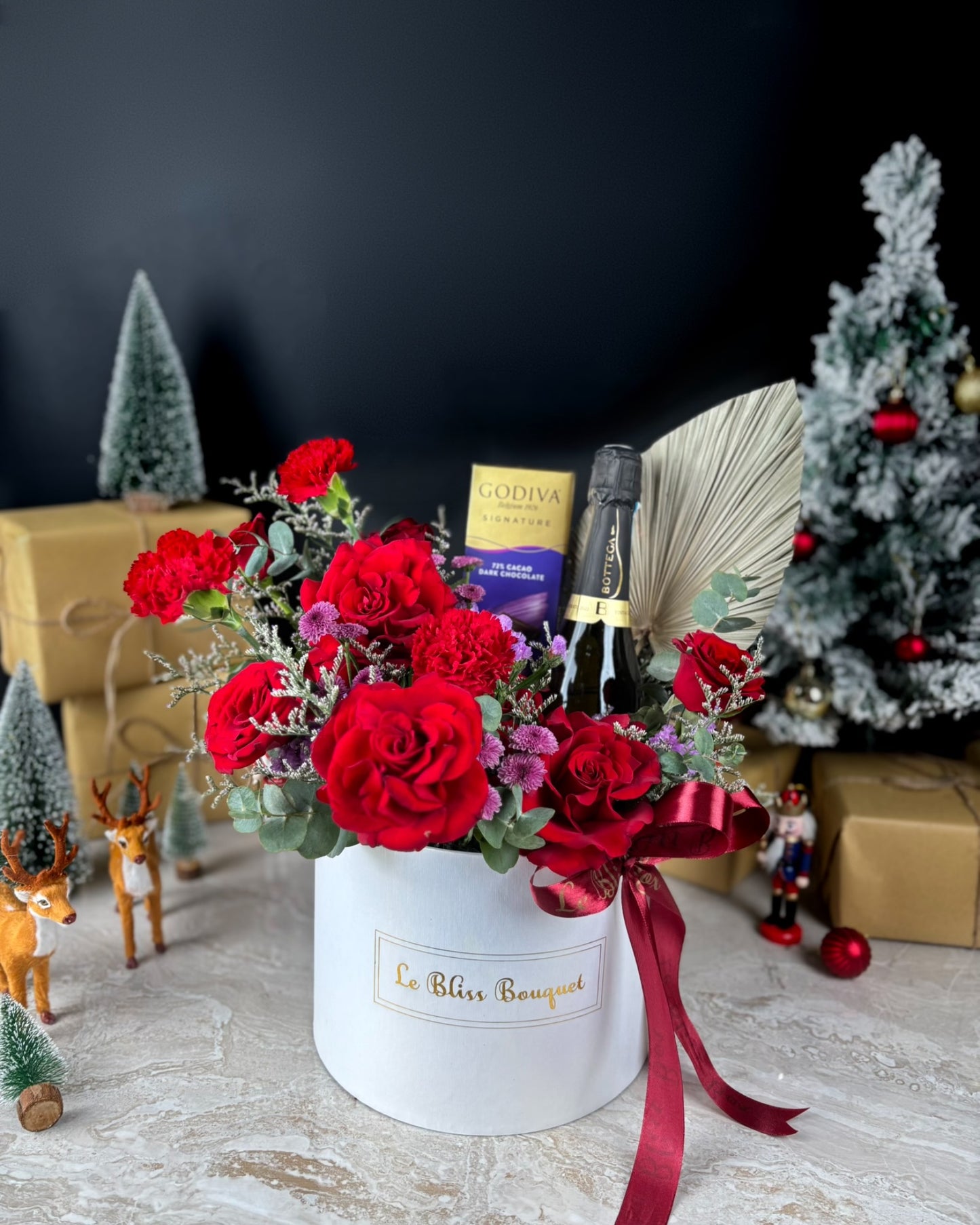 Bloombox of red roses in a white box with a bottle of champagne and Godiva chocolate bars in the background. Discover the best Christmas hamper in Jakarta & Surabaya. The Bottega Godiva Bloombox by Le Bliss Box blends luxury wine, chocolates & fresh blooms.