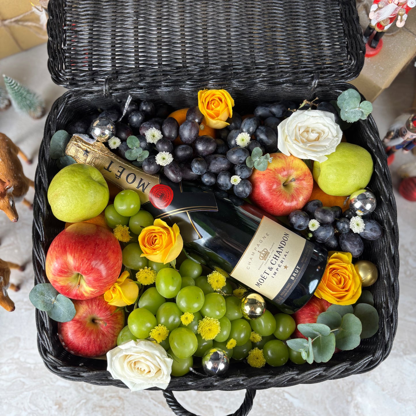 Wine hamper Surabaya with fruits, flowers, and a bottle of Moet & Chandon champagne. Fruit basket with moet champagne bottle, apples, grapes, and flowers on a textured surface. Midnight Moët Champagne Hamper | Best Christmas Hamper in Jakarta, Surabaya & Tangerang – Le Bliss Box