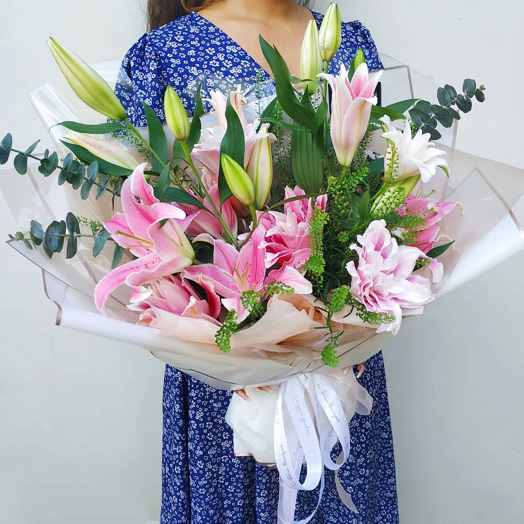 A person holding a large bouquet of pink casablanca lilies wrapped in white paper with a ribbon. Le Bliss Bouquet’s Double Lily Pink Large Bouquet — soft pink lilies, elegant wrapping, perfect for gifting. Delivery in Jabodetabek, Surabaya & Cirebon.