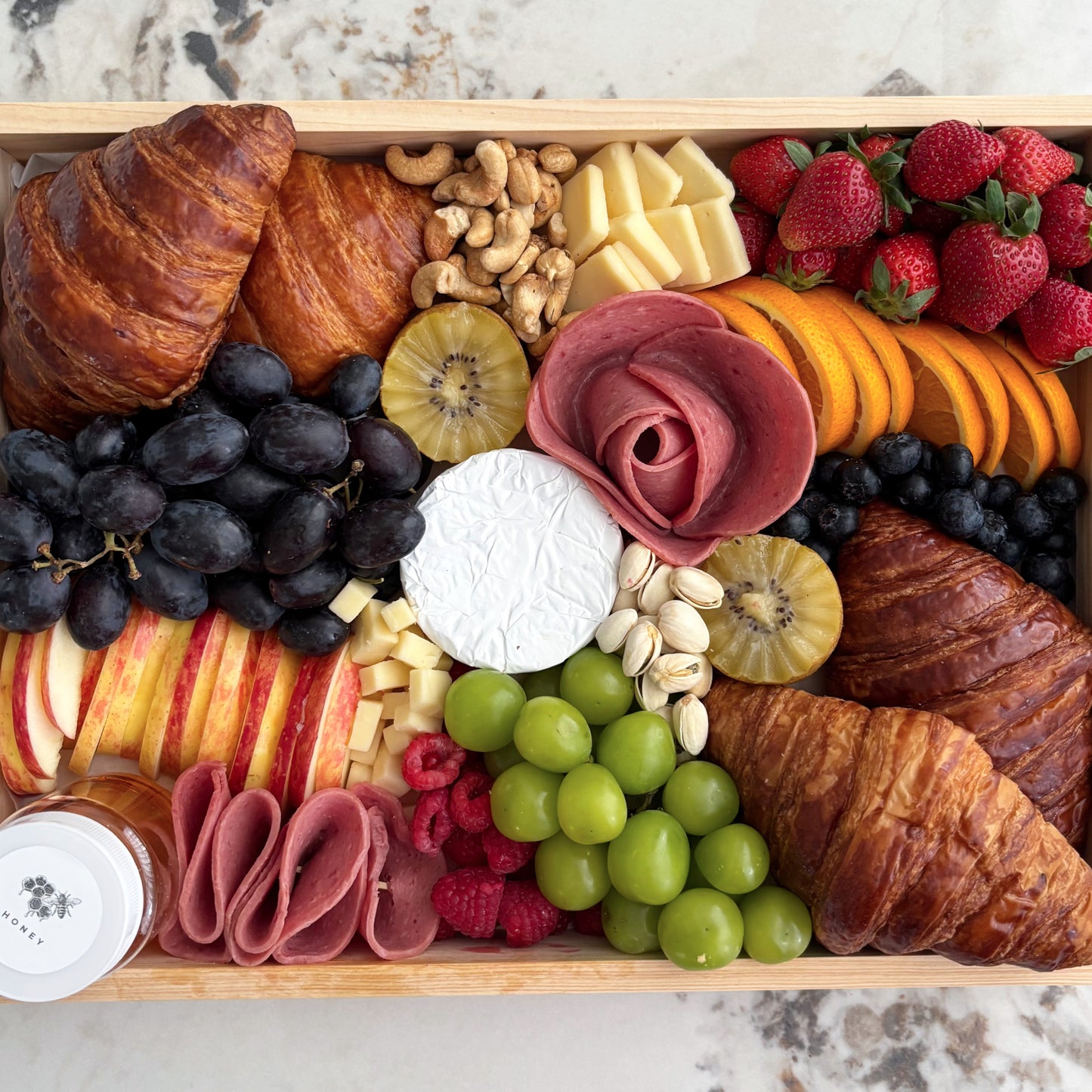 Assorted food platter with fruits, meats, and cheeses on a wooden board. Wooden box filled with assorted fruits, cheeses, and pastries on a marble surface. Brunch Croissant Cheese Platter | Best Cheese Platter in Jakarta & Surabaya – Le Bliss Platter