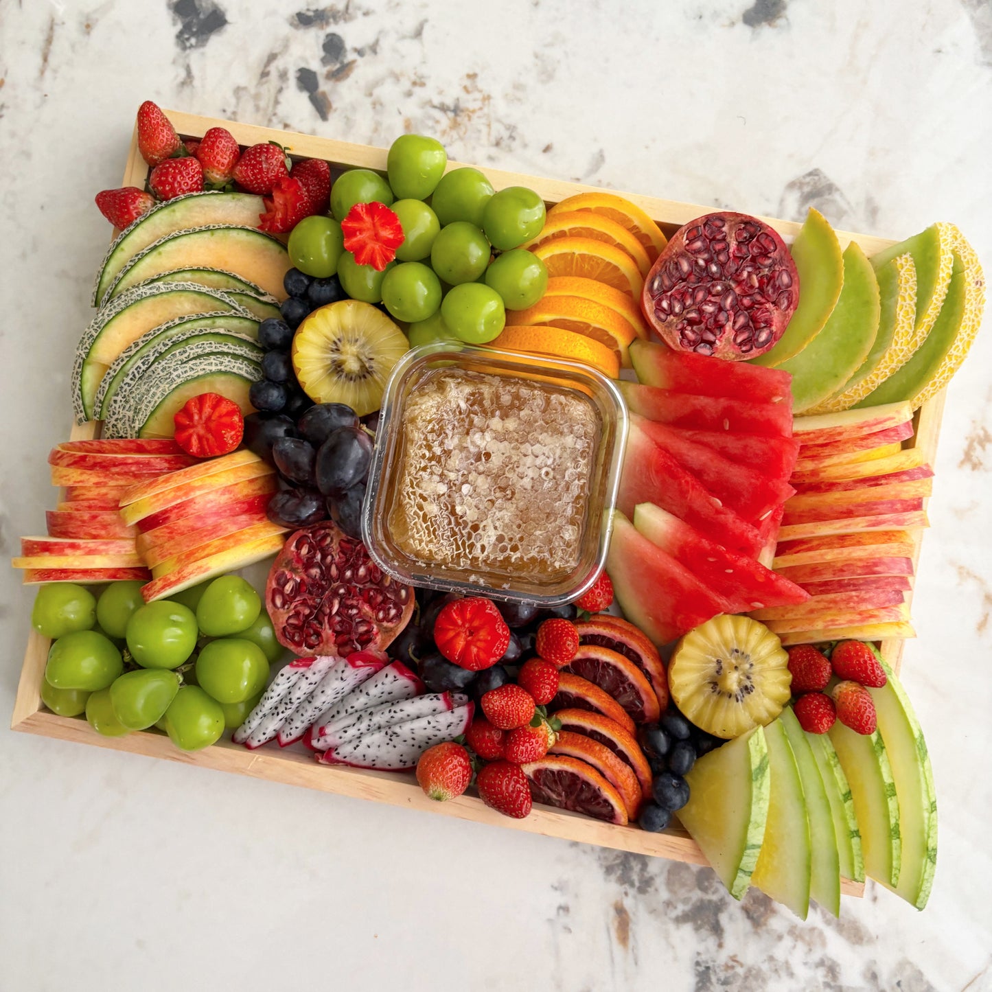 Assorted fruit platter with a central bowl of honey on a marble surface. Le Bliss Platter is part of Le Bliss, offering premium cheese, fruit, charcuterie & sweet platters across Jakarta, Surabaya & Tangerang.