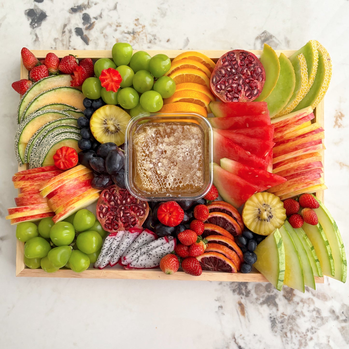 Assorted fruit platter with a central container of honey on a marble surface. Honeycomb Colorful Fruit Platter | Fresh Luxury Fruit Platter Jakarta & Surabaya – Le Bliss Platter