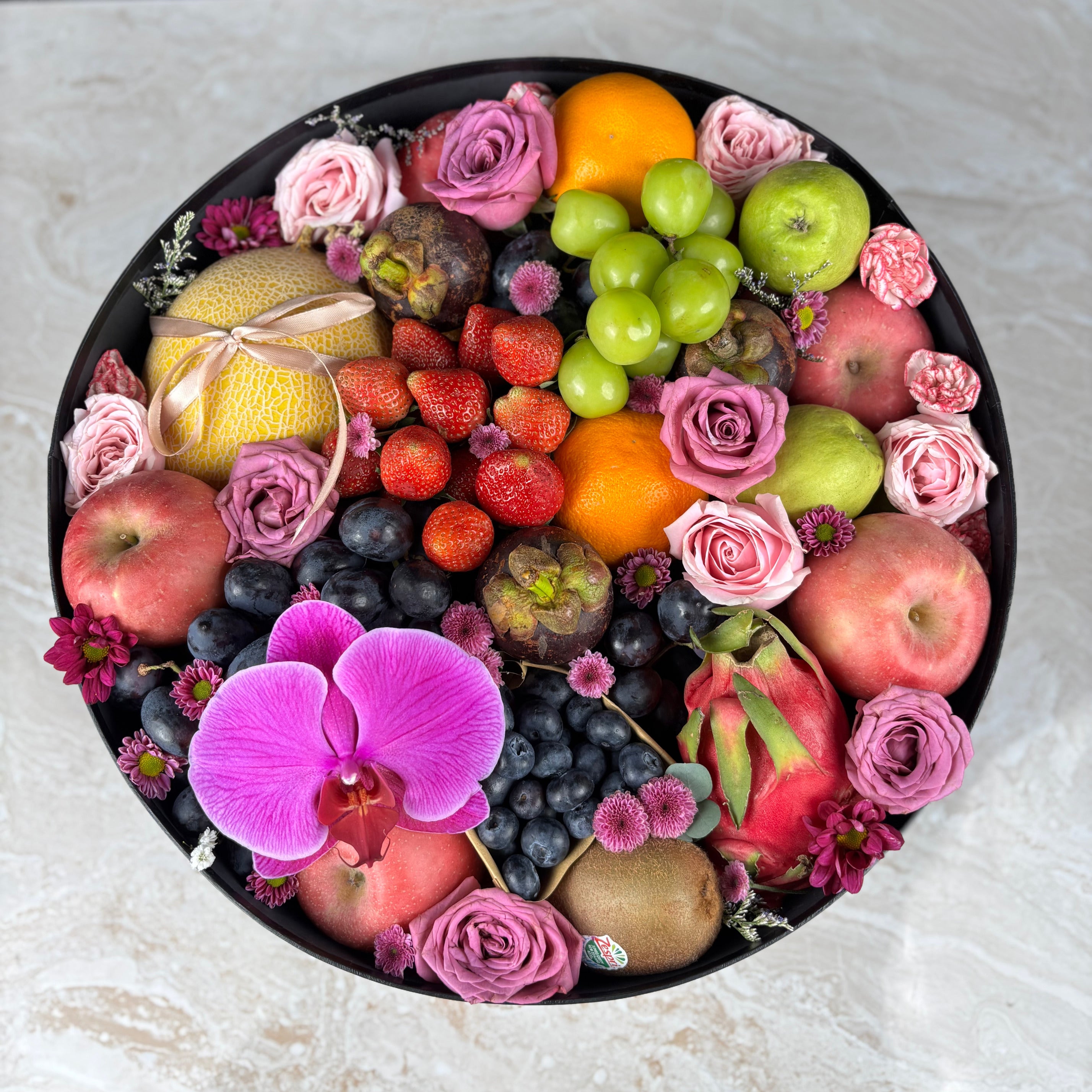 Assorted fruits and flowers arranged in a circular pattern on a white surface. Fruit and flower arrangement branding. Parcel Buah Surabaya, Jakarta, Cirebon.