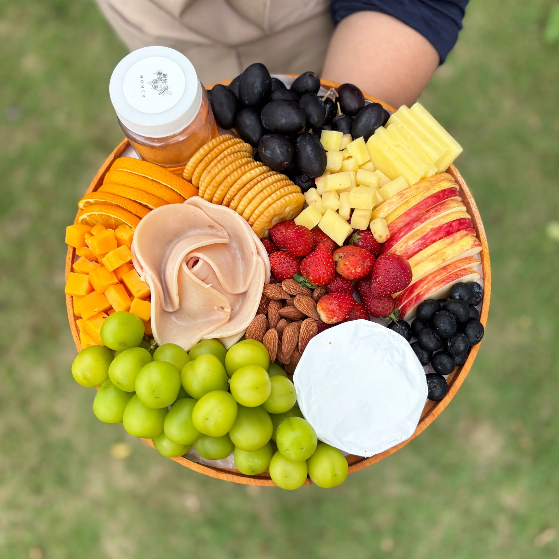 Assorted fruits, cheeses, and crackers on a platter held by a person outdoors. chicken salami cheese platter. Assorted fruits, cheeses, and nuts on a platter with a blurred green background. best cheese platter in jakarta, surabaya. 
