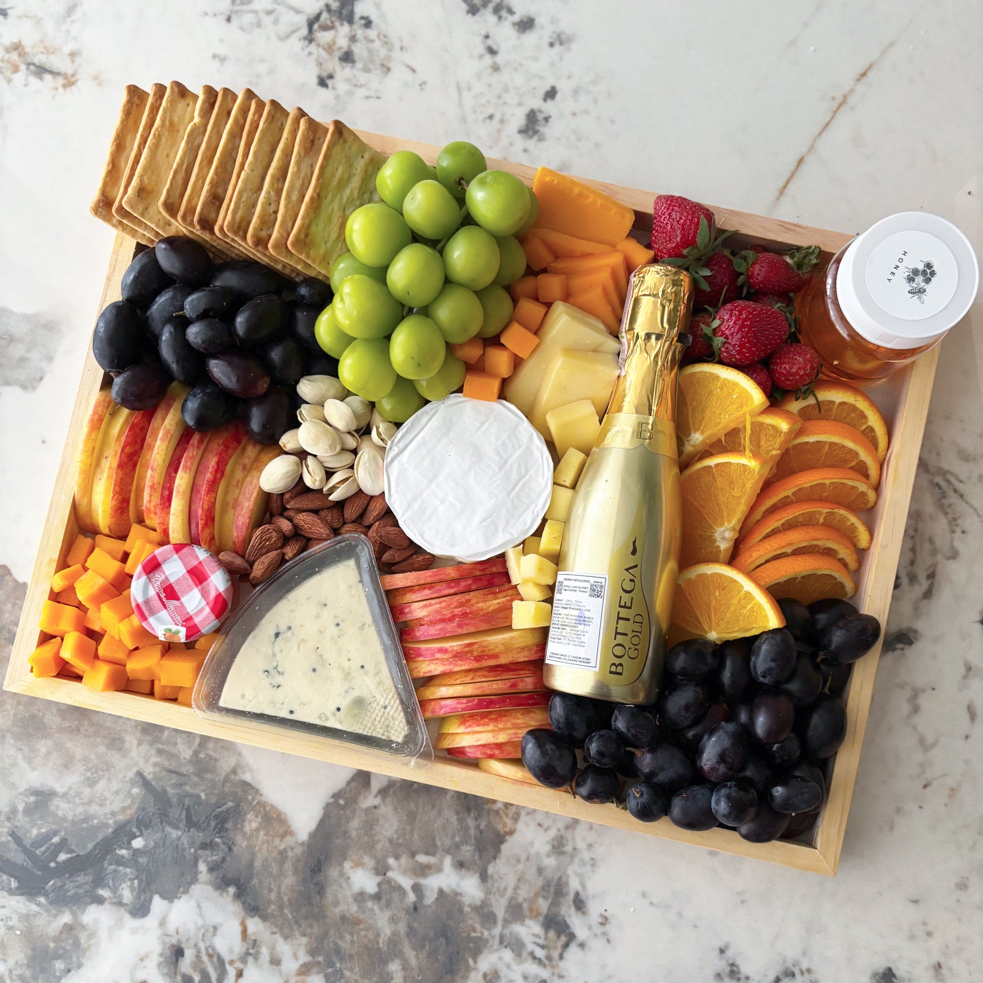 Assorted fruits, cheeses, and crackers on a wooden board with a bottle of champagne. Bottega Wine Cheese Platter | Best Wine & Cheese Platter in Jakarta & Surabaya – Le Bliss Platter
