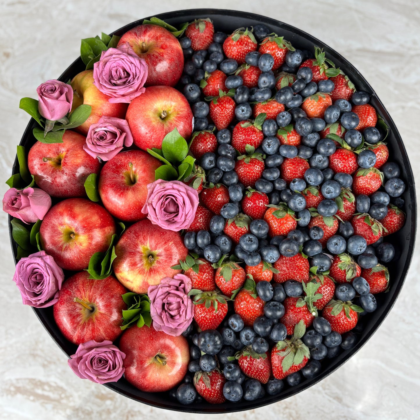 Assorted fruits including apples, strawberries, and blueberries arranged in a circular pattern on a black plate with pink flowers. Fruit and flower arrangement in a black box with 'Le Bliss Box' branding. Decorative box filled with fruits and flowers, including apples, strawberries, and blueberries, with a 'Happy Mother's Day' card. kado hari ibu surabaya, jakarta, cirebon