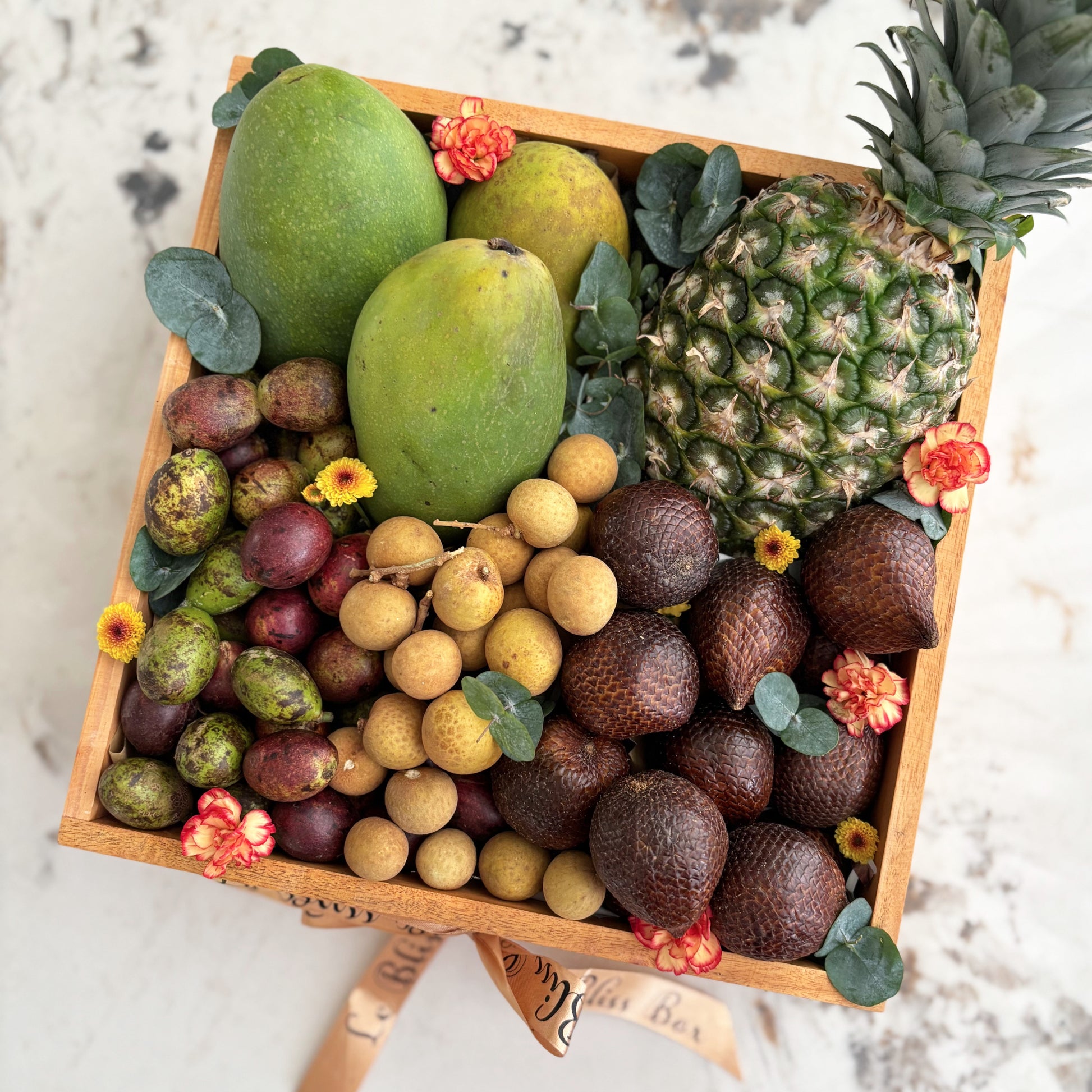 Assorted fruits including mangoes, pineapples, and longans in a wooden box on a marble surface. tropical fruits hamper jakarta, surabaya, cirebon