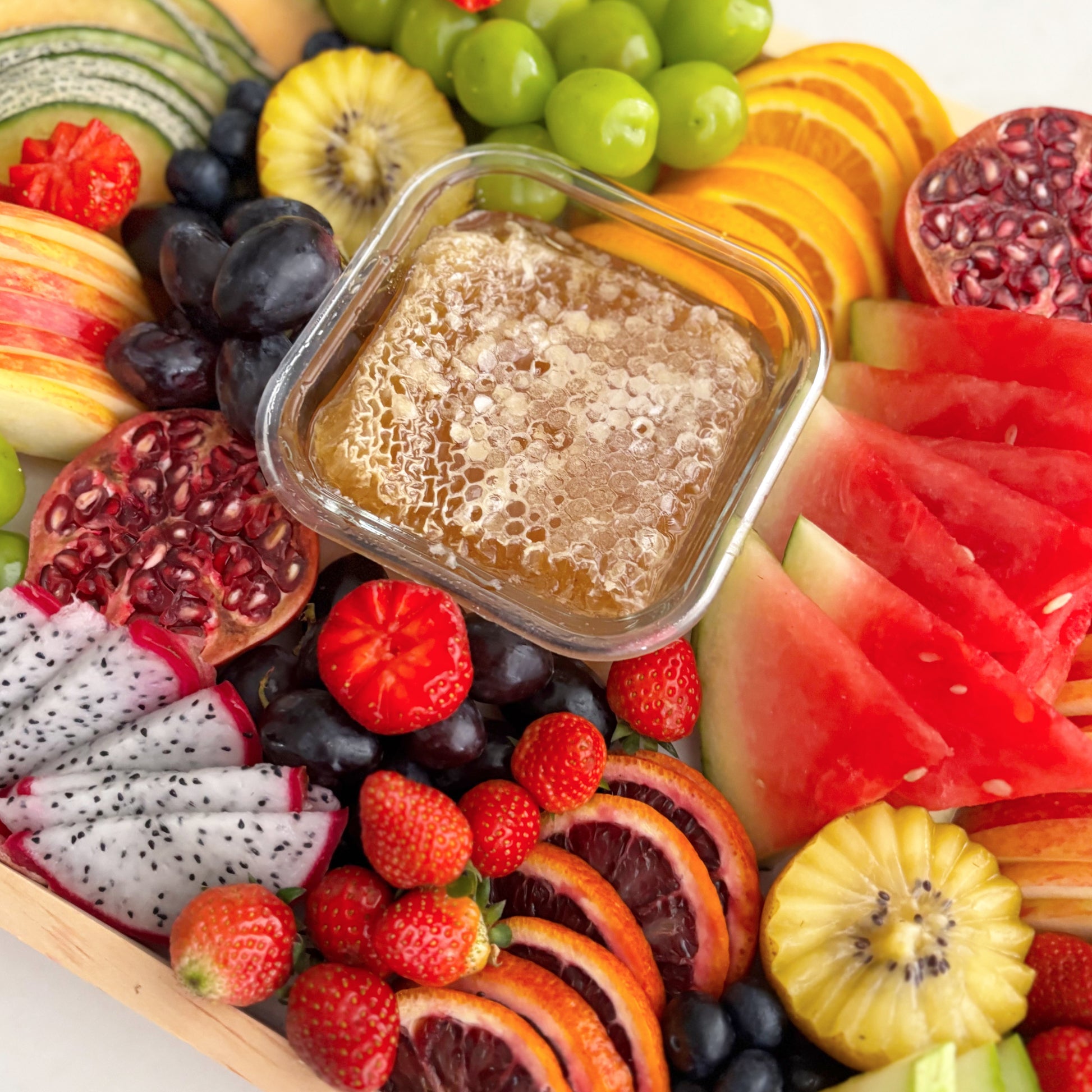 Assorted fruits including watermelon, dragon fruit, and kiwis with a container of honey on a wooden board. Le Bliss Platter