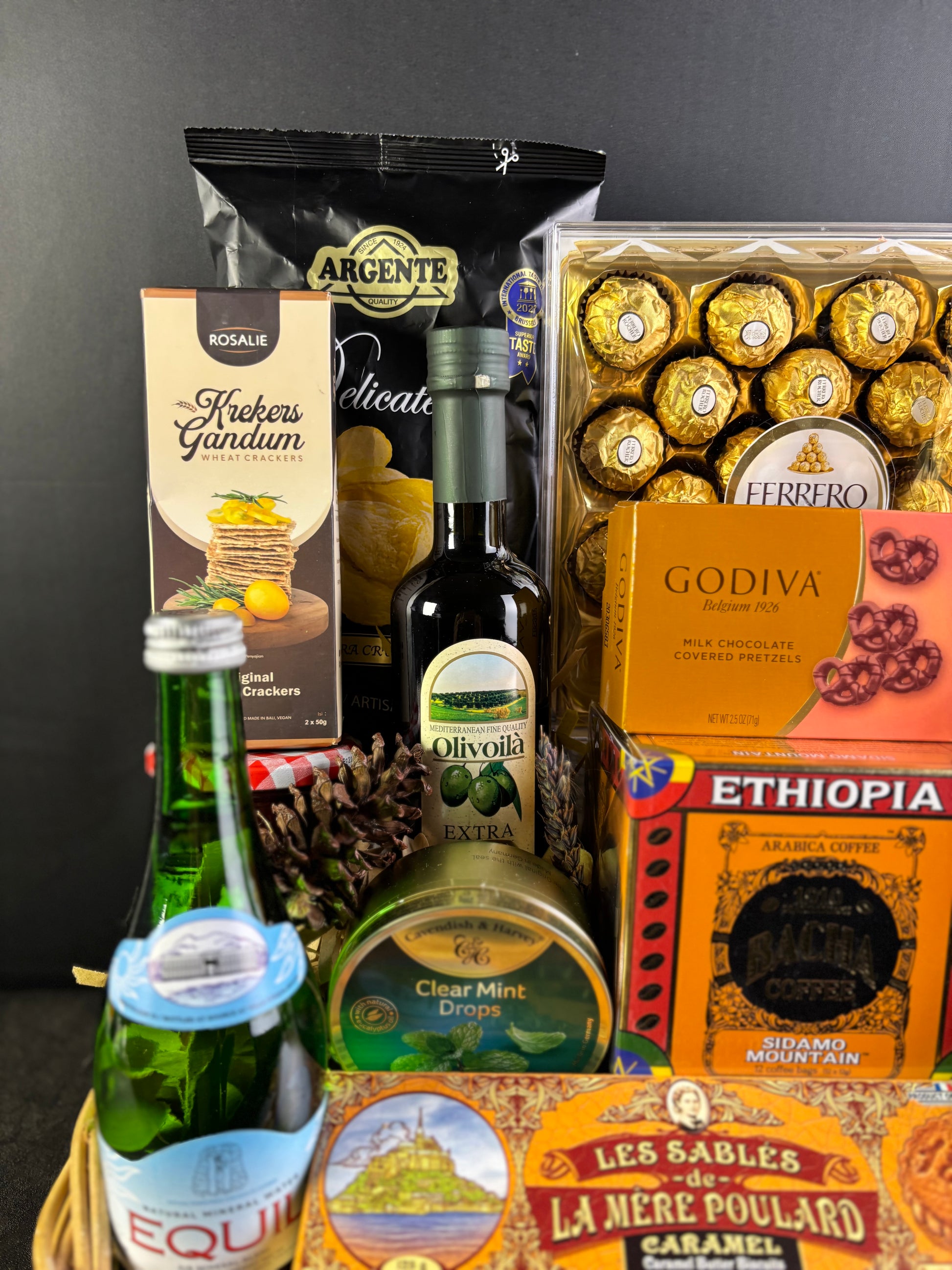 Assorted gourmet items including chocolate, olive oil, and candy on a dark background. basket with snacks, drinks, and chocolates. hamper jakarta, hamper surabaya, gourmet hamper jakarta. hamper by le bliss box