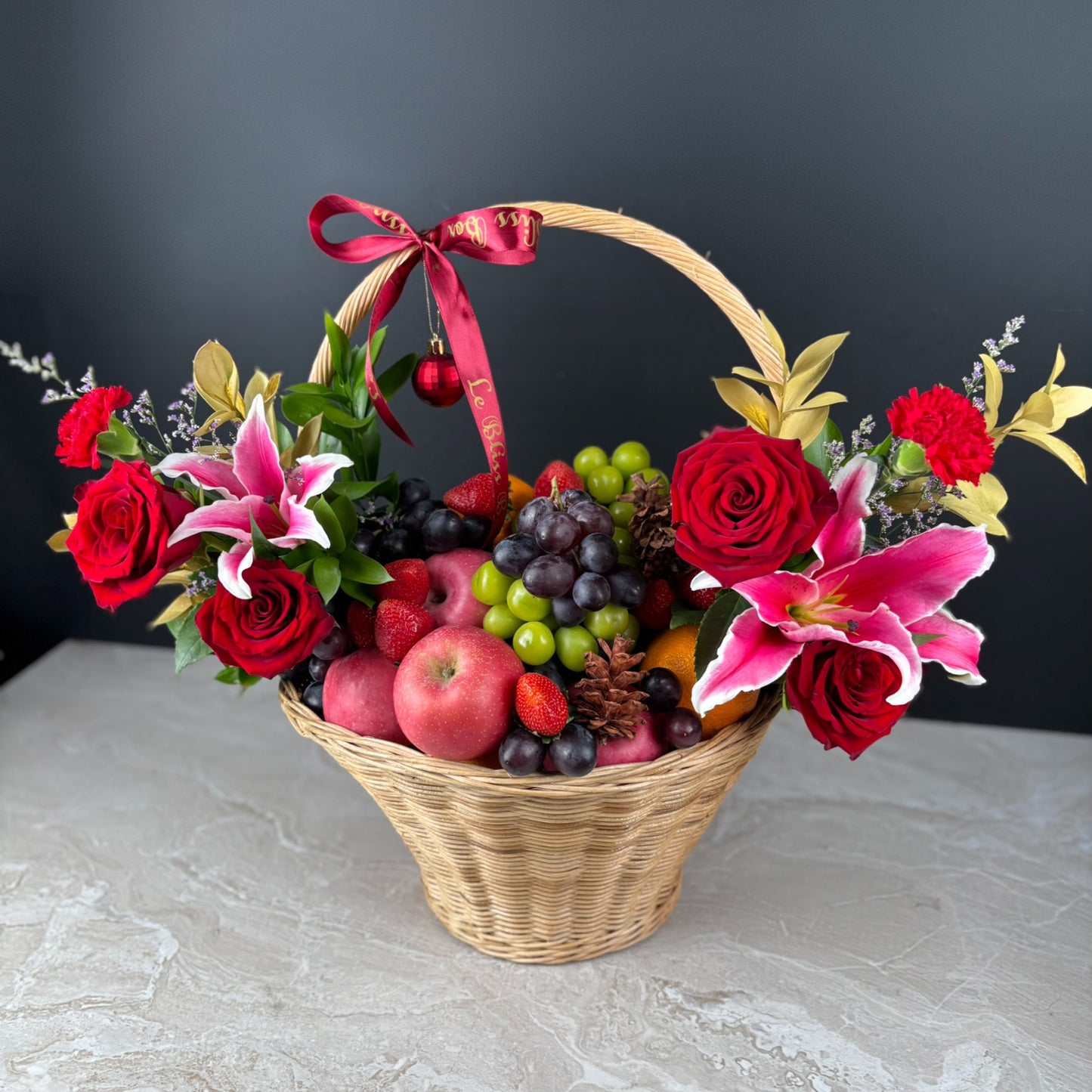 Basket of flowers and fruits with a red ribbon on a dark background. Celebrate the season with the Festive Royal Harvest Fruity Parcel by Le Bliss Box. Premium imported fruits beautifully styled for gifting in Jakarta, Tangerang, Cirebon & Surabaya.