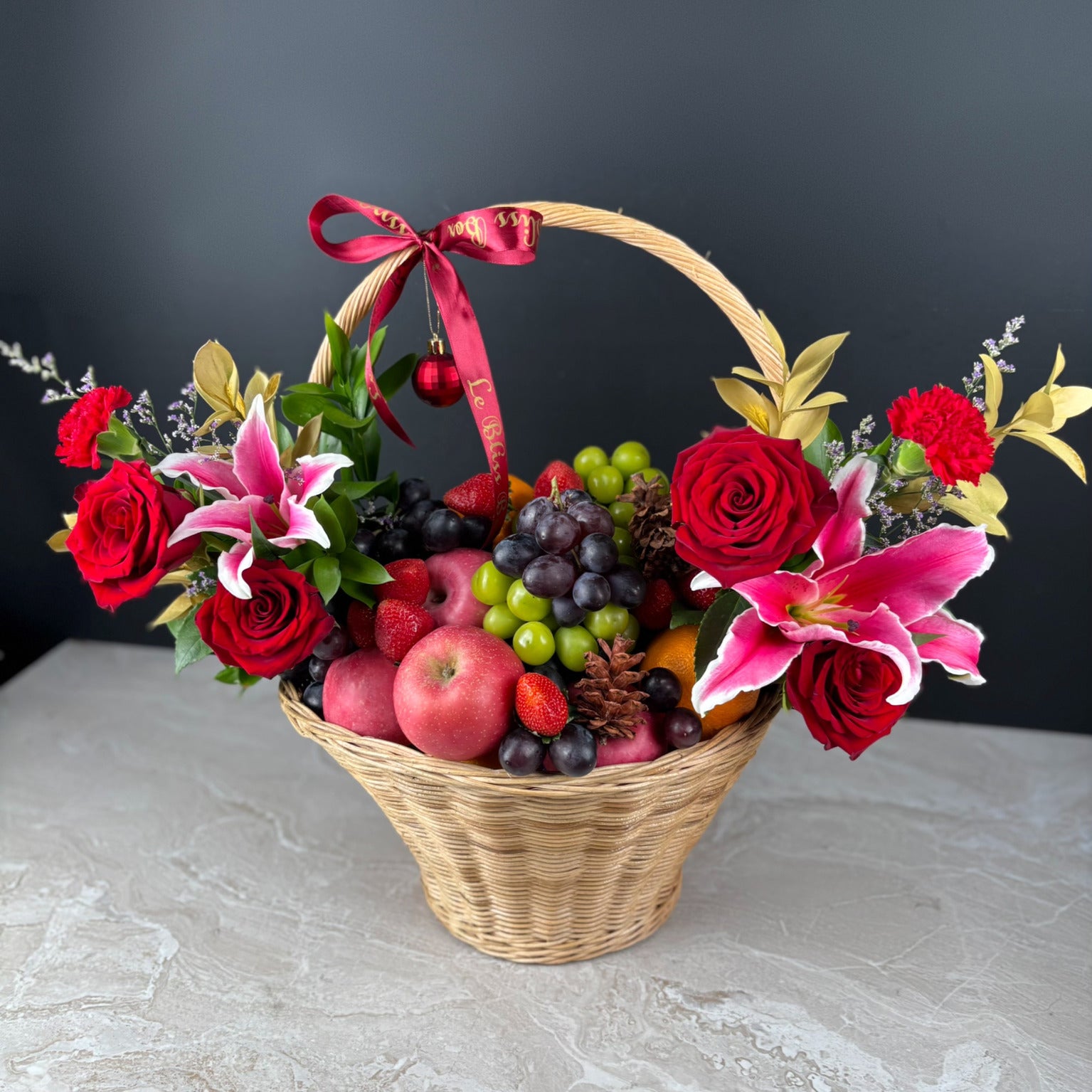 Basket of flowers and fruits with a red ribbon on a dark background. Celebrate the season with the Festive Royal Harvest Fruity Parcel by Le Bliss Box. Premium imported fruits beautifully styled for gifting in Jakarta, Tangerang, Cirebon & Surabaya.