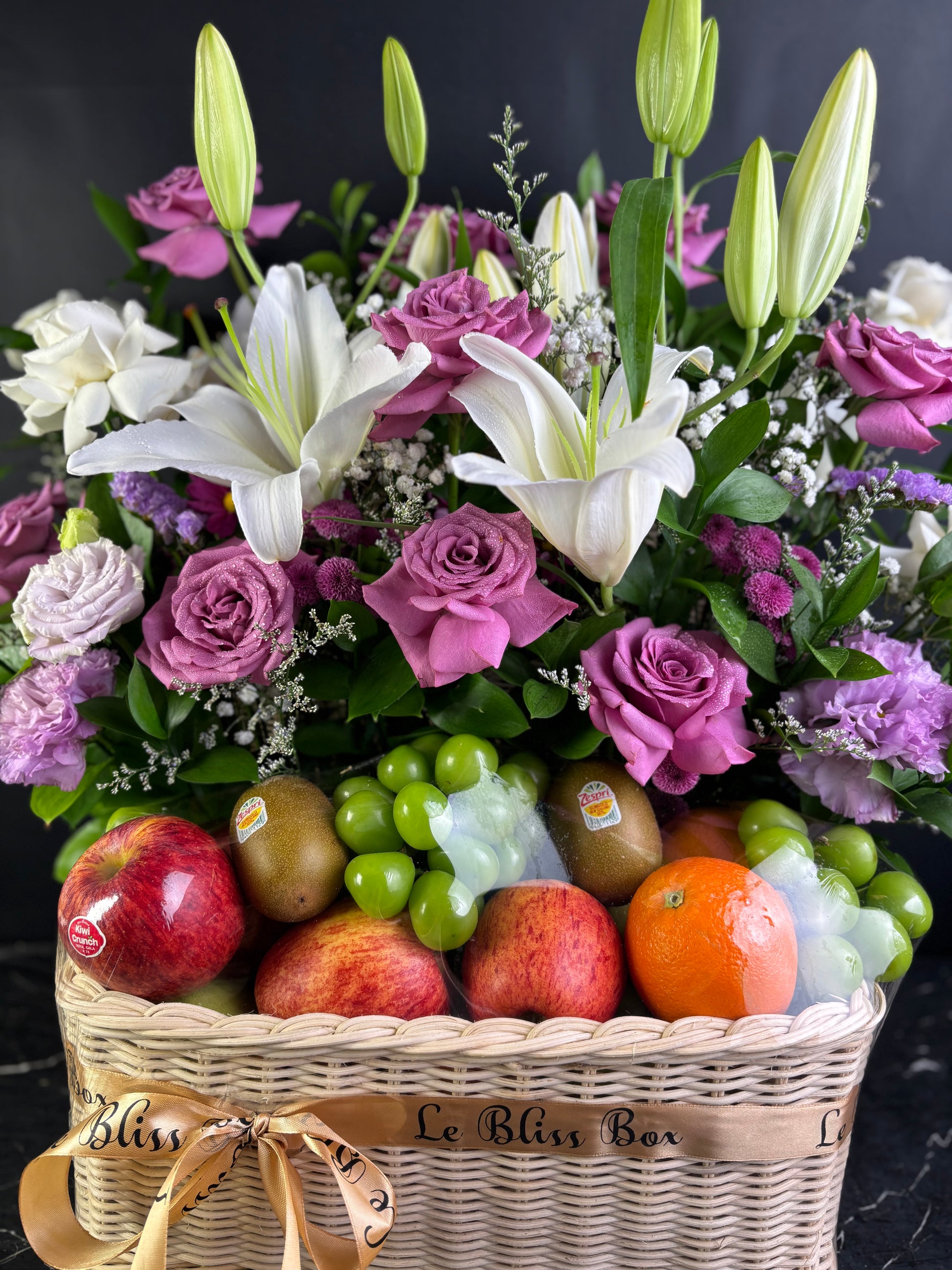Basket of flowers with apples, grapes, and oranges on a dark background. Basket of purple flowers and fruits on a dark surface with a black background. Fruit hamper jakarta, cirebon, surabaya, tangerang. by le bliss box, parcel buah premium terbaik