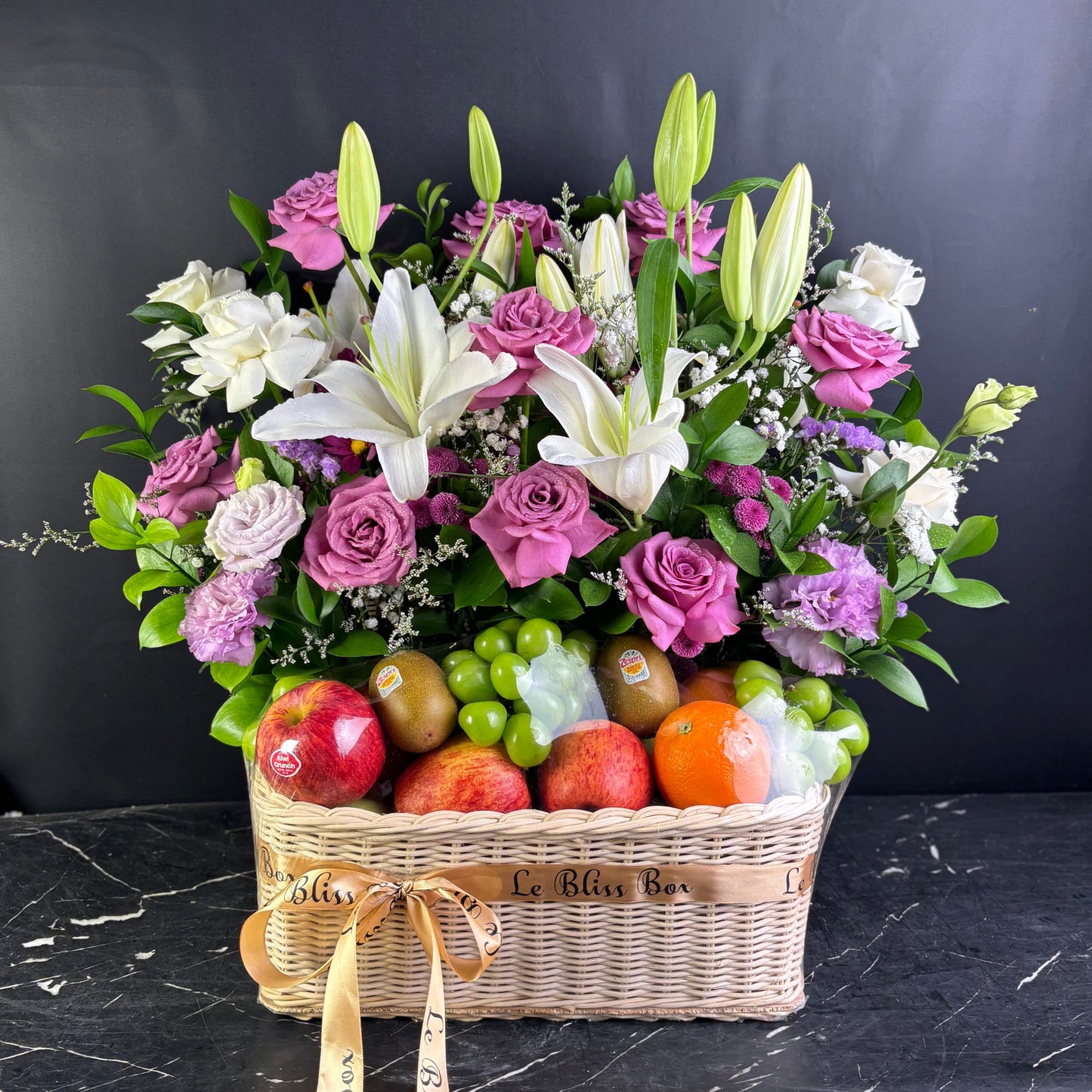 Basket of purple flowers and fruits on a dark surface with a black background. Fruit hamper jakarta, cirebon, surabaya, tangerang. by le bliss box, parcel buah premium terbaik