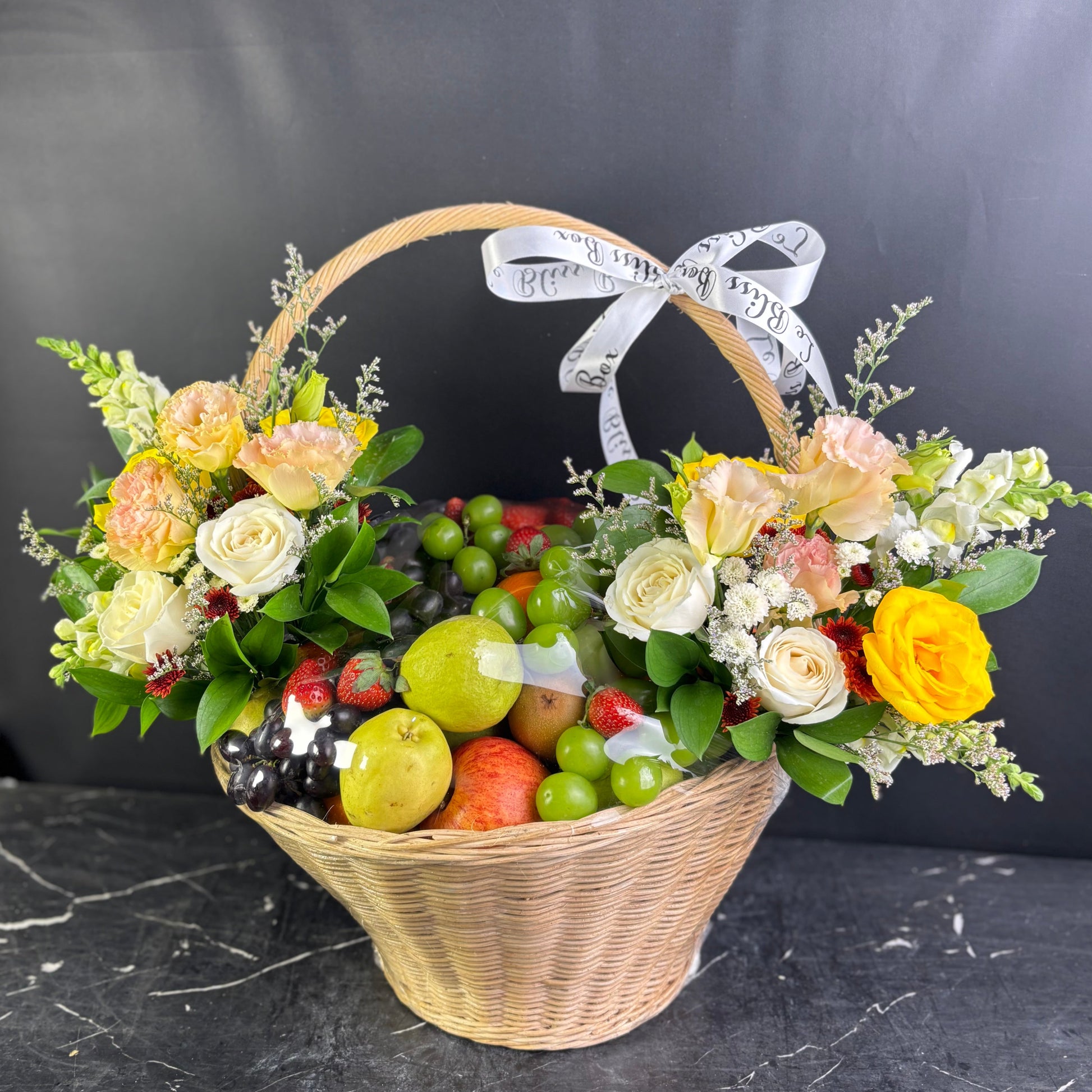 Basket with flowers and fruits on a dark surface. Royal harvest fruity parcel, parcel buah surabaya, parcel buah jakarta, parcel buah indonesia, fruit hamper delivery by le bliss box. parcel buah bunga gresik, tangerang, sidoarjo