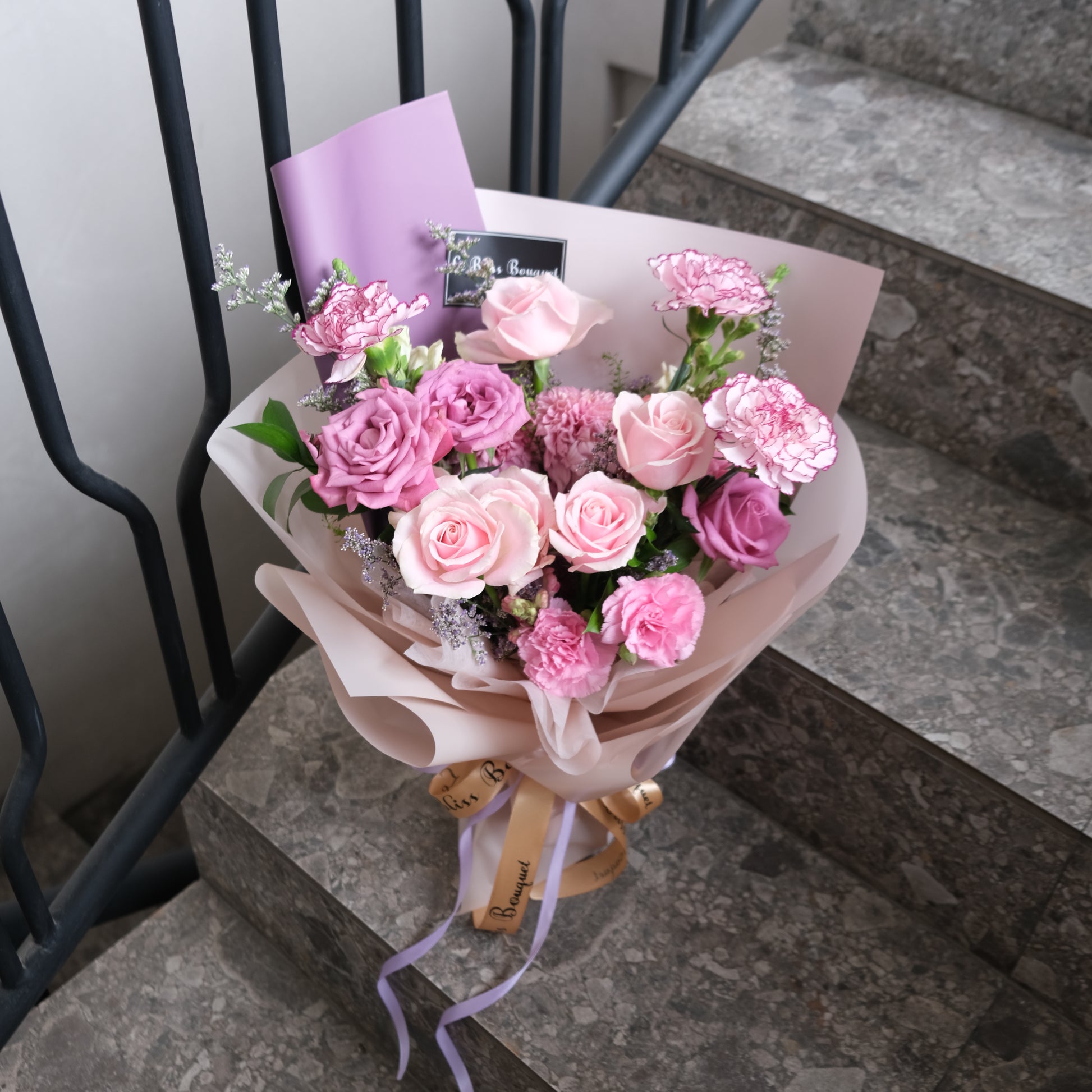 Bouquet of pink flowers with a card on a stone surface. Medium Bouquet dari Le Bliss Bouquet. Kado Hari Ibu yang elegan dengan mawar pink & lilac. Pengiriman Surabaya, Jakarta & Cirebon. Premium & heartfelt.