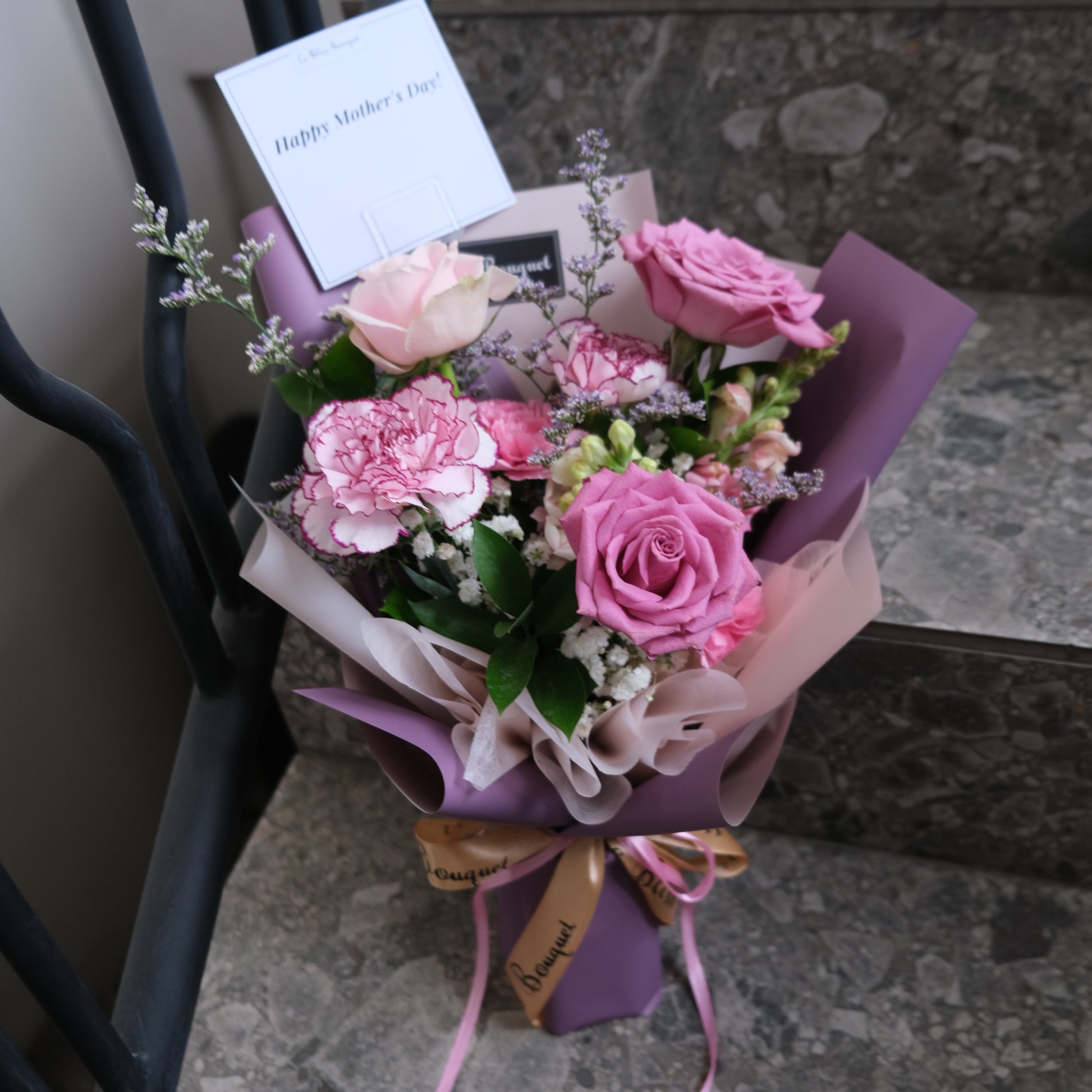 Bouquet of pink flowers with a card on a stone surface. Pink Lilac Sonata Smallie Bouquet dari Le Bliss Bouquet. Kado Hari Ibu elegan dengan bunga pink & lilac. Pengiriman Surabaya, Jakarta & Cirebon. Fresh, sweet, premium.