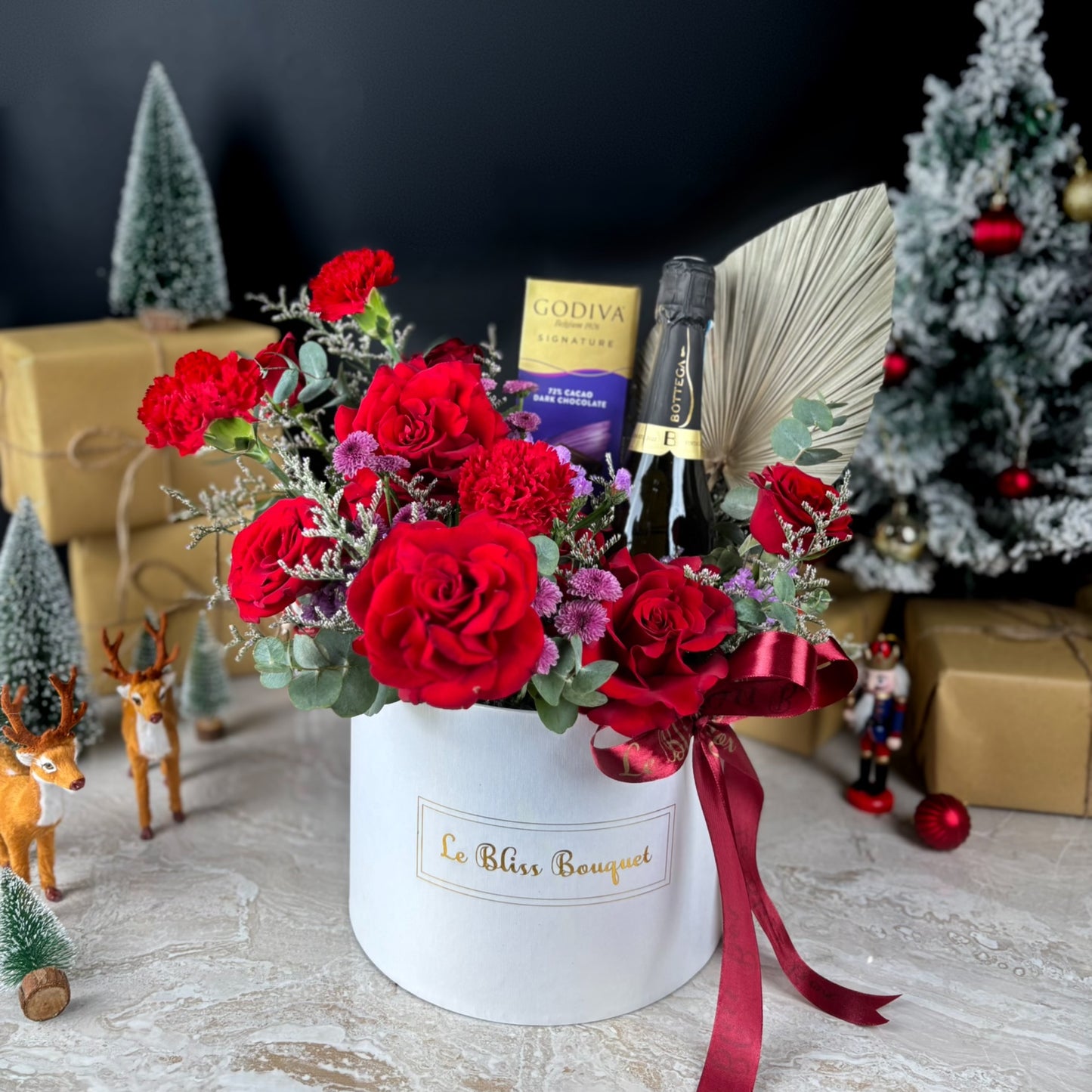 Bouquet of red roses in a white box with a bottle of champagne and Godiva chocolate bars in the background. Discover the best Christmas hamper in Jakarta & Surabaya. The Bottega Godiva Bloombox by Le Bliss Box blends luxury wine, chocolates & fresh blooms.