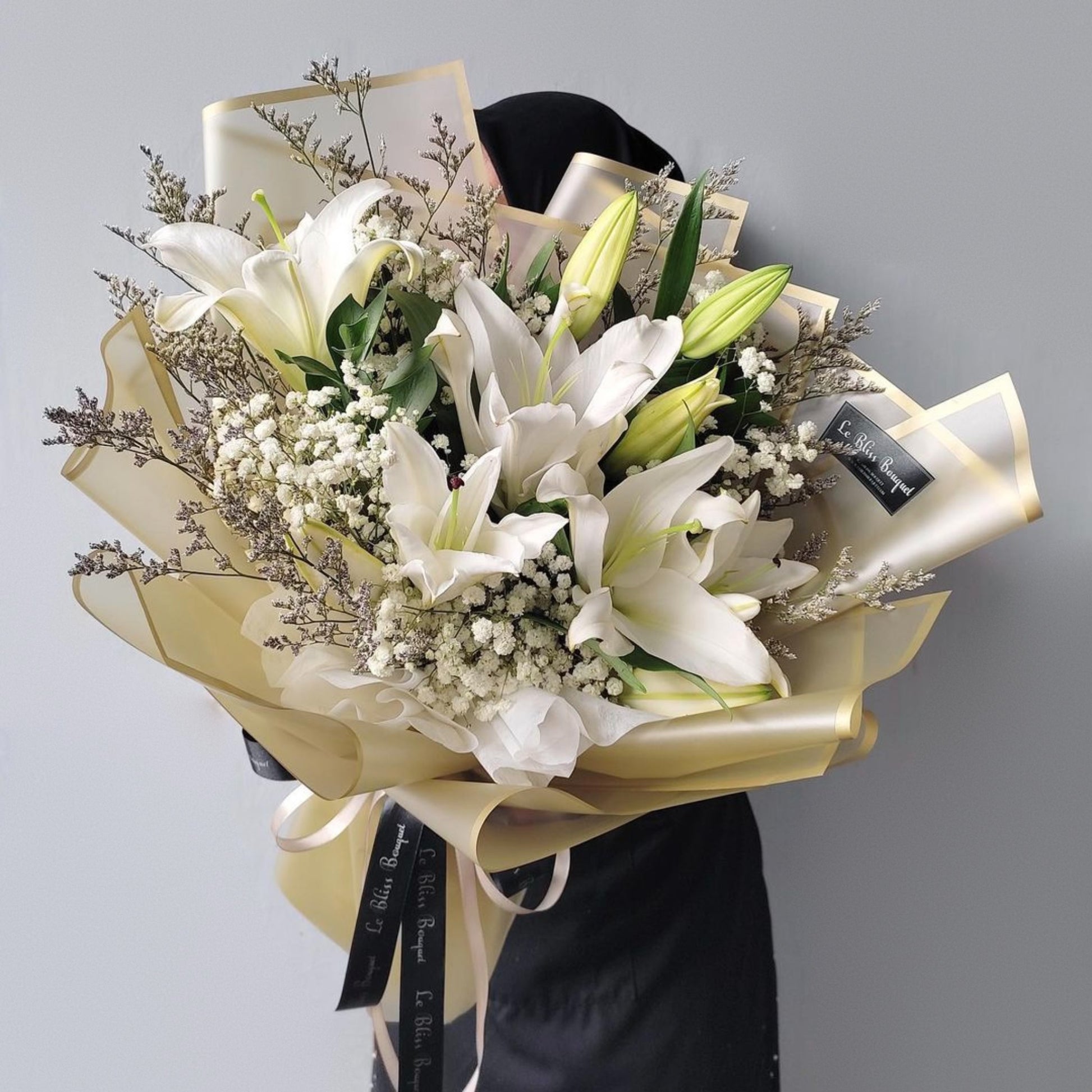 Bouquet of white lilies and greenery with a yellow ribbon against a gray background. Le Bliss Bouquet’s White Lily Large Bouquet is perfect for love, sympathy, or celebration. Delivery to Jabodetabek, Surabaya & Cirebon.
