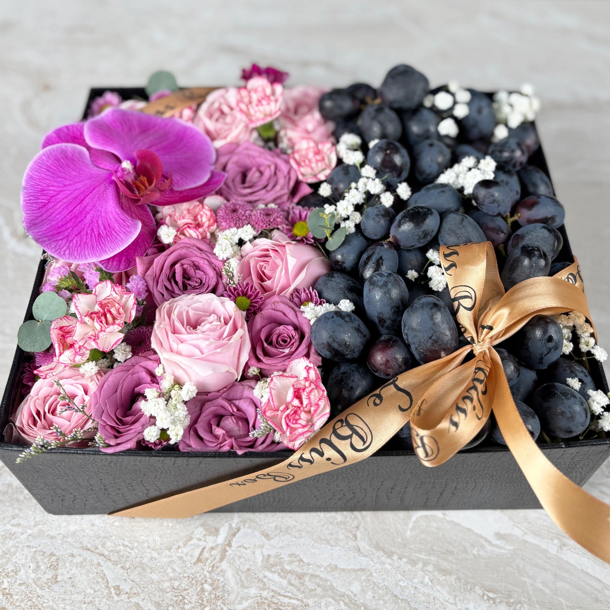 Box of flowers and grapes with a gold ribbon on a light background. Pink Lilac Sonata Autumn Grape Box dari Le Bliss Bouquet. Kado Hari Ibu elegan dengan bunga & buah premium. Pengiriman Surabaya, Jakarta & Cirebon. Soft, sweet & luxurious. Kado Anggur  dan anggrek untuk Hari Ibu