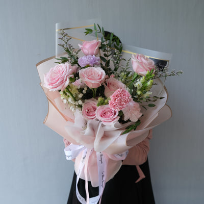A medium-sized bouquet of pink flowers with green leaves, wrapped in a pink and white paper with a ribbon. florist surabaya,  toko bunga surabaya, bouquet surabaya, buket bunga surabaya