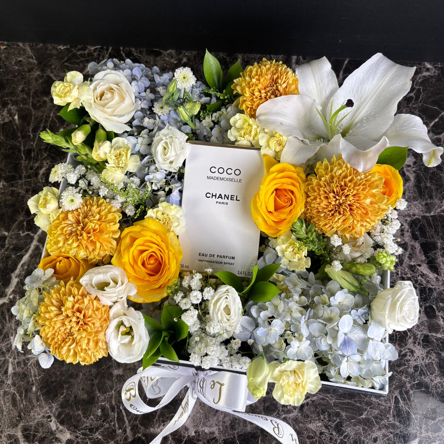 Floral arrangement with a Chanel perfume bottle surrounded by yellow and white flowers on a dark background. chanel perfume hamper by le bliss bouquet.