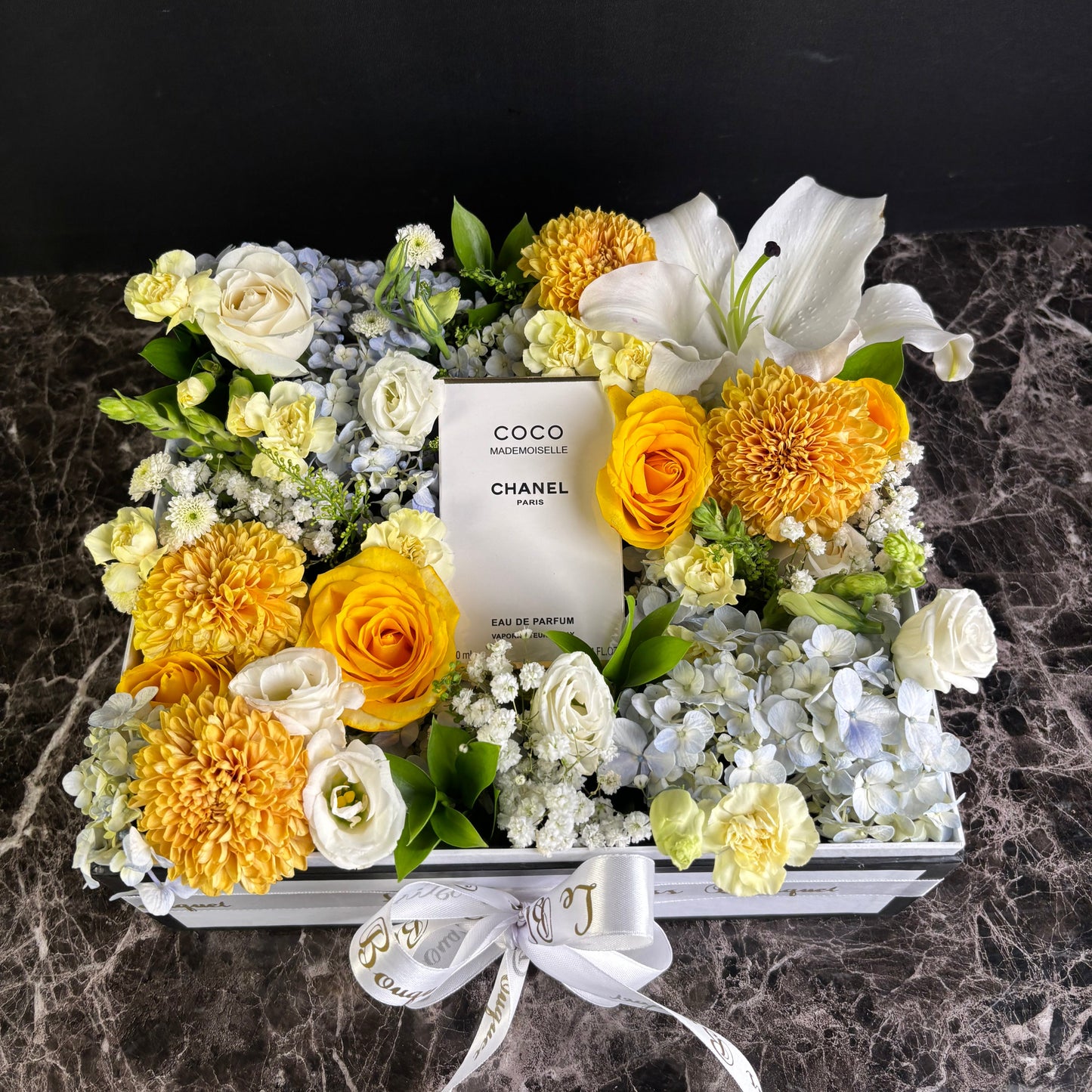 Floral arrangement with a Chanel perfume bottle surrounded by yellow and white flowers on a dark background. chanel perfume hamper by le bliss bouquet