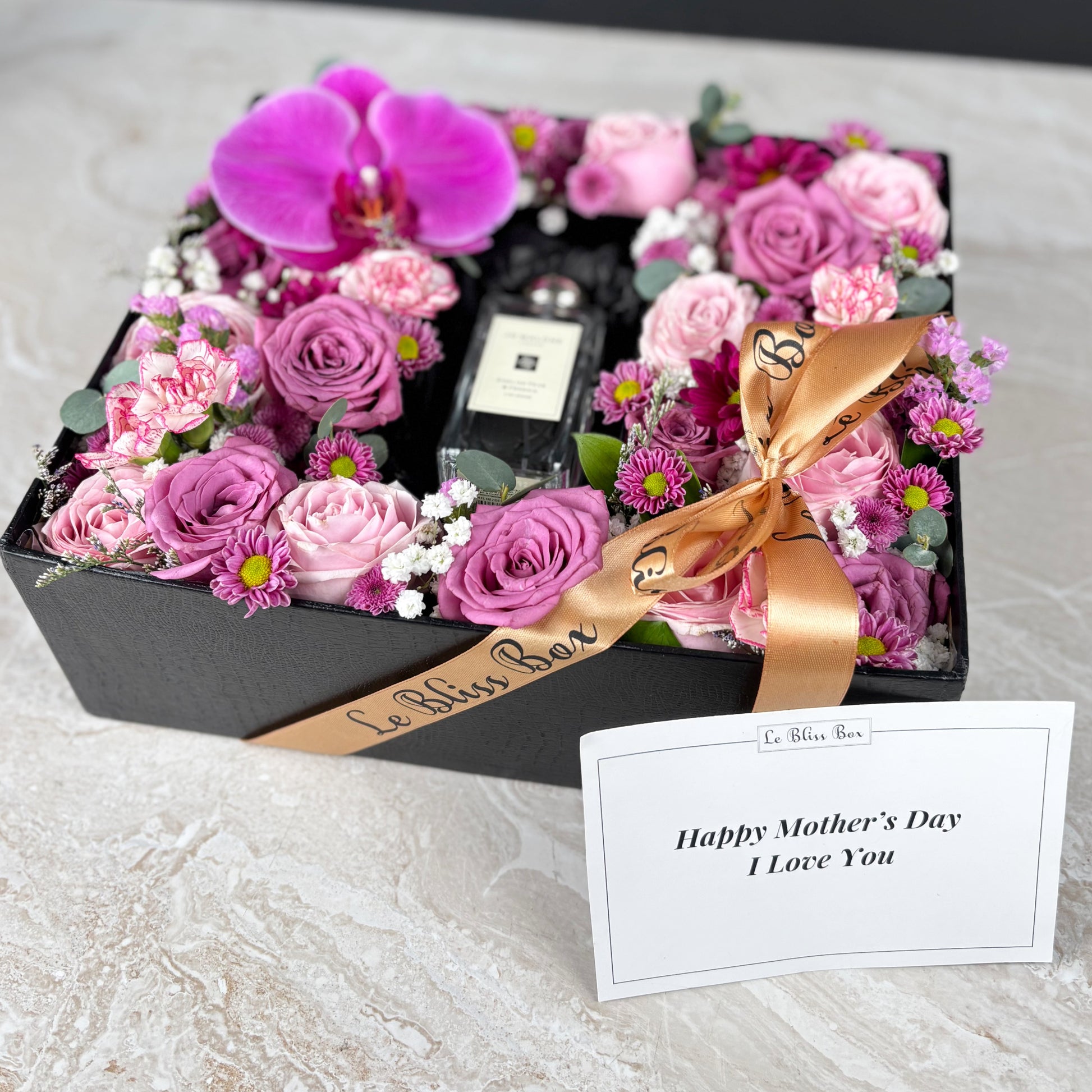 Floral arrangement with a bottle of perfume and a card on a textured surface. Kado Hari Ibu Surabaya, Kado Hari Ibu Jakarta