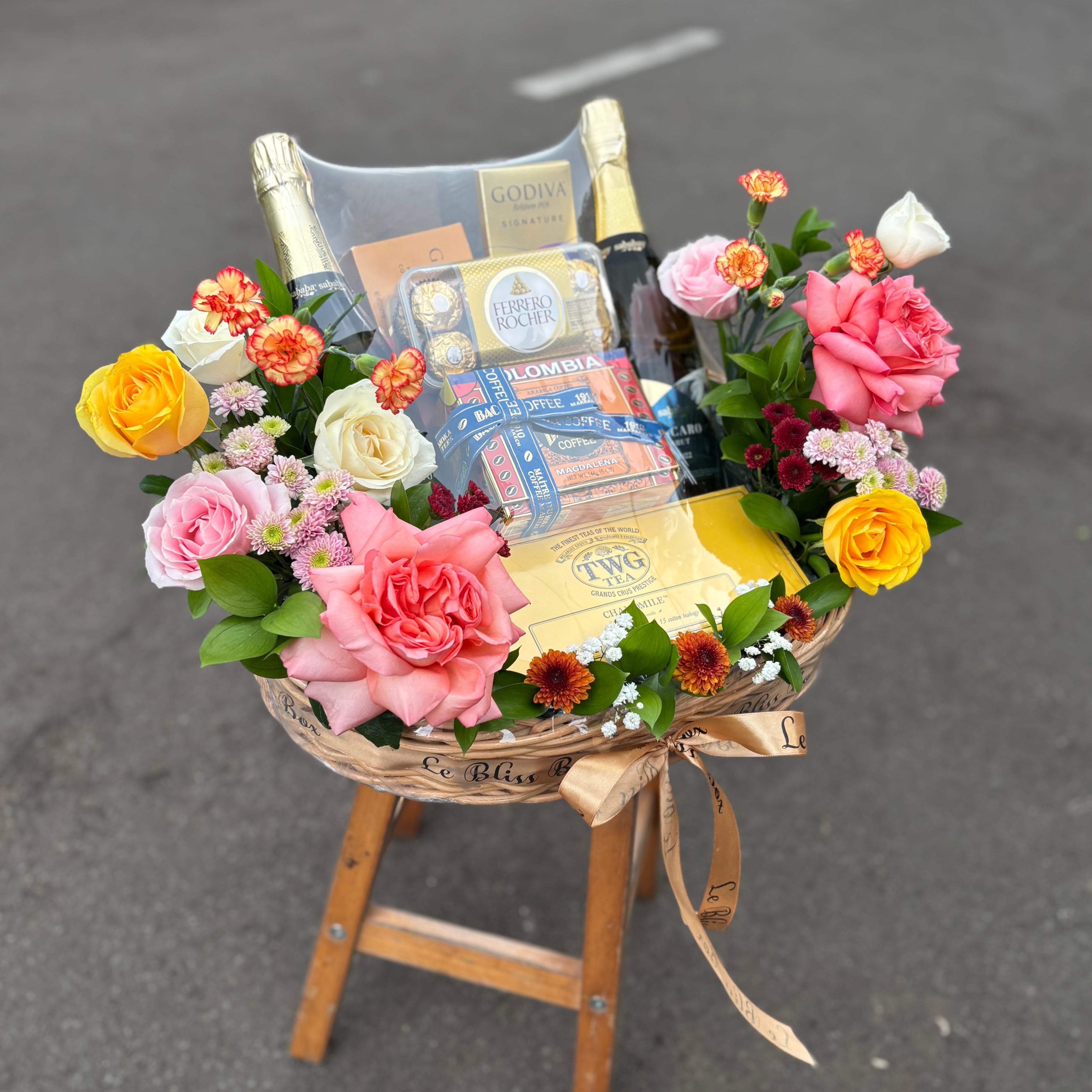 Floral arrangement with a bottle of wine and gift items on a small wooden stool outdoors. gourmet hamper jakarta, gourmet hamper surabaya, gourmet hamper tangerang, TWG Tea, bacha coffee, ferrero, sababay wine with fresh flowers by le bliss