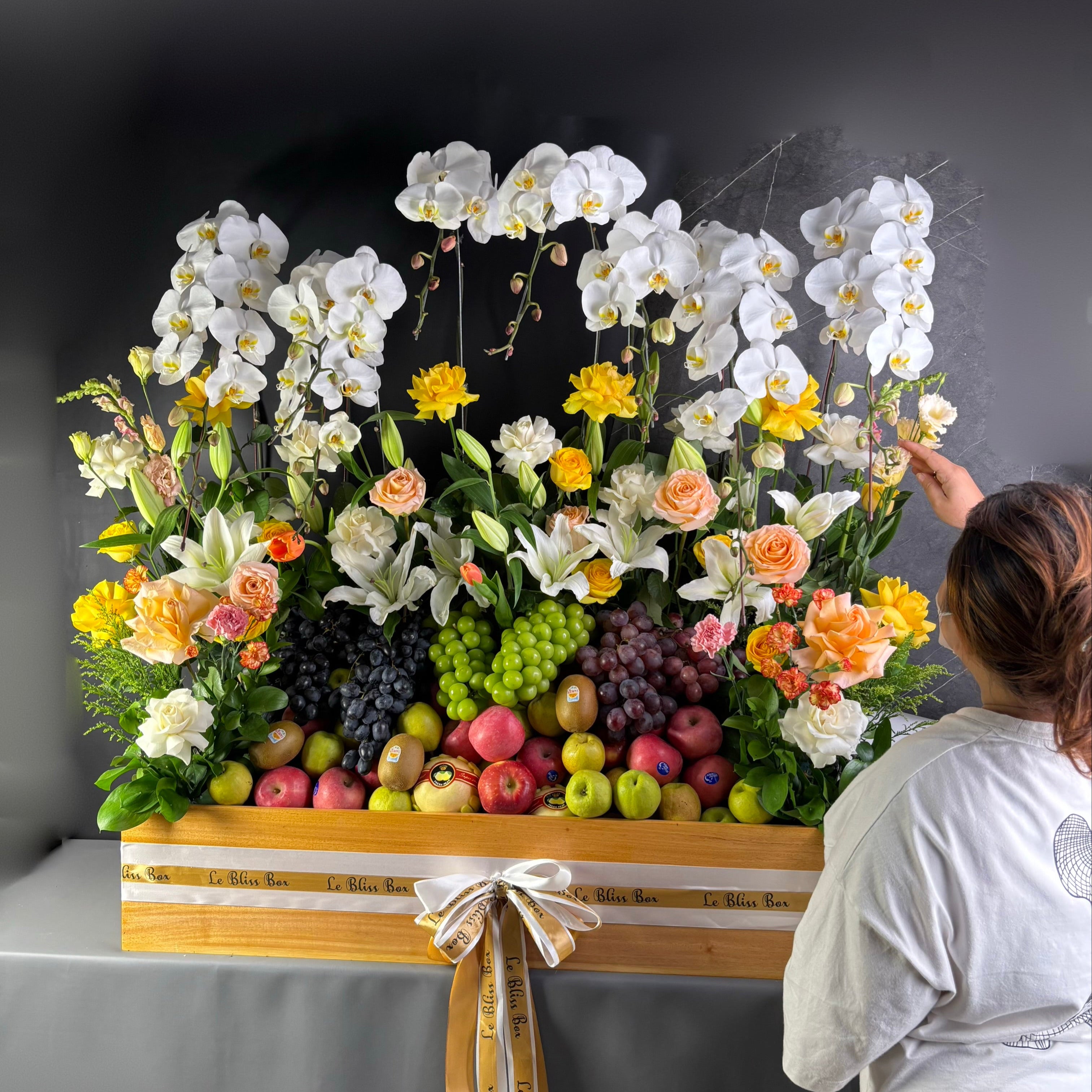 Floral arrangement with fruits on a table, person adjusting flowers. Floral arrangement with fruits on a wooden stand against a dark background. rangkaian bunga dan parcel buah besar jakarta. by le bliss bouquet