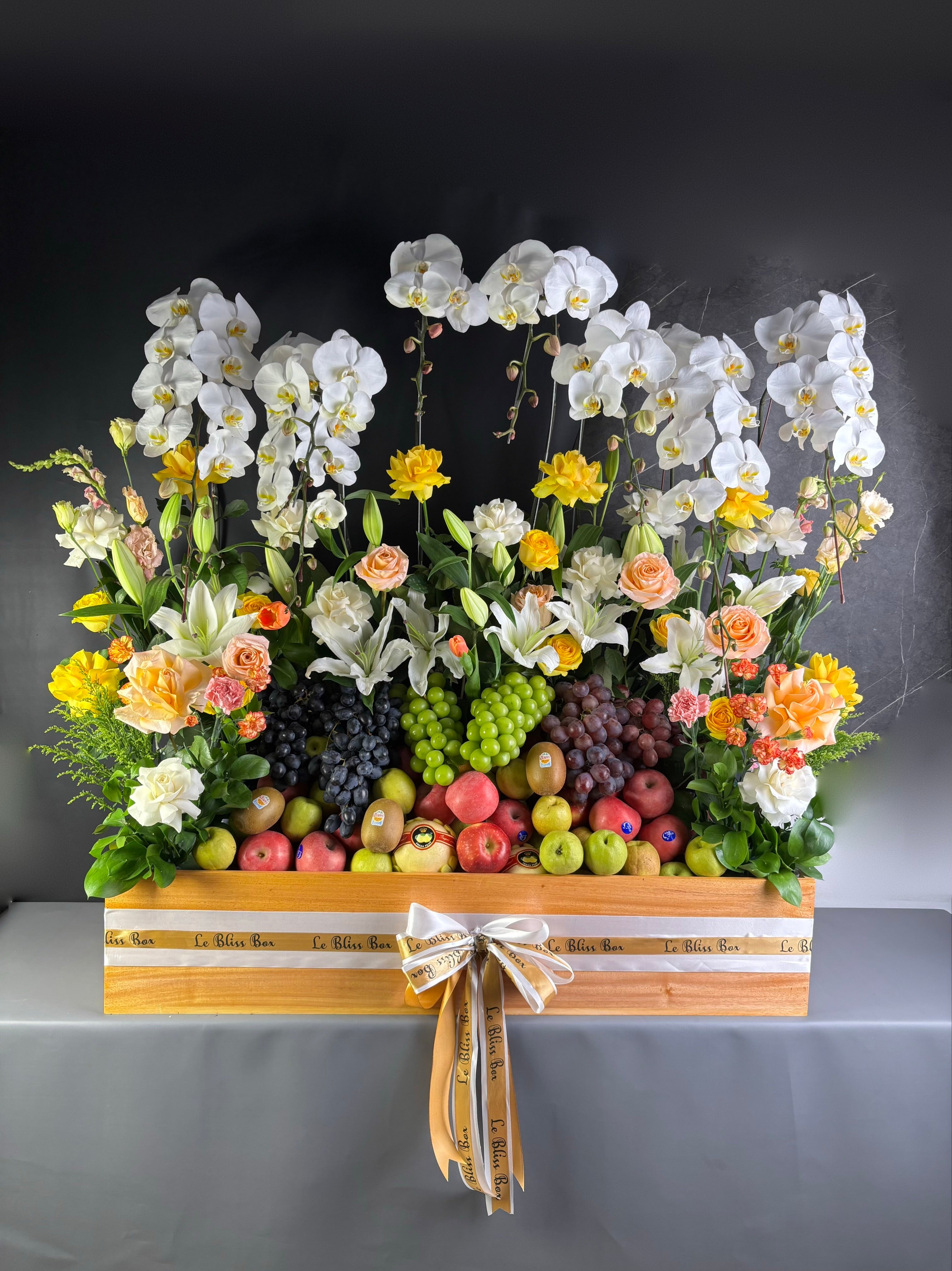 Floral arrangement with fruits on a wooden stand against a dark background. rangkaian bunga dan parcel buah besar jakarta. by le bliss bouquet