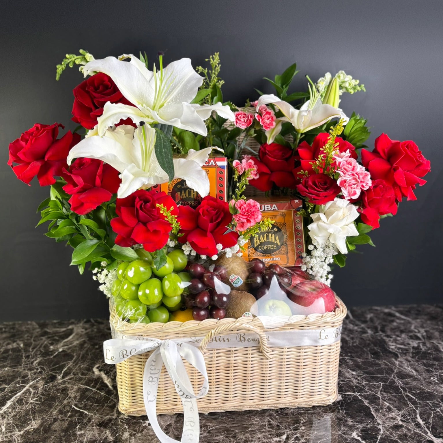 Floral arrangement with red and white flowers, fruits, and a box of bacha coffee in a woven basket on a dark surface. kado hari ibu surabaya, kado surabaya 