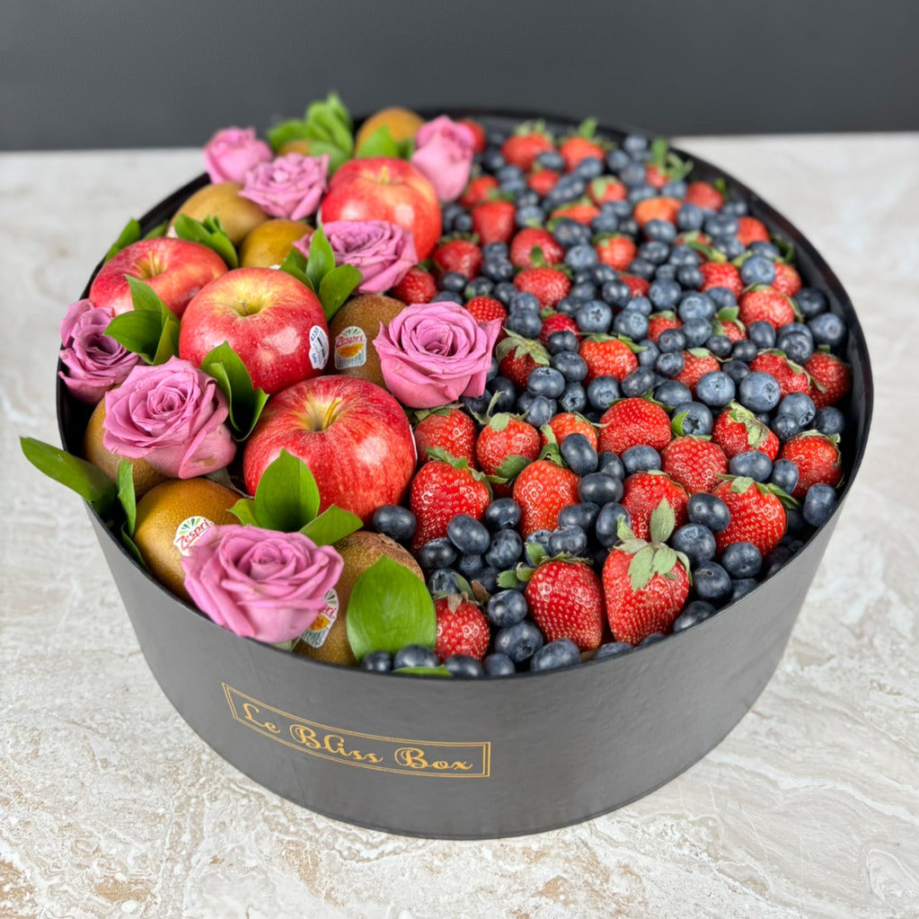 Fruit and flower arrangement in a black box labeled 'Le Bliss Box' on a light surface.Decorative box filled with fruits and flowers on a light surface. Premium Apple, Kiwi & Berries Round Fruit Box dari Le Bliss Box. Parcel buah segar untuk hadiah mewah di Surabaya, Jakarta, Tangerang & Cirebon. Fresh, elegant, and healthy.
