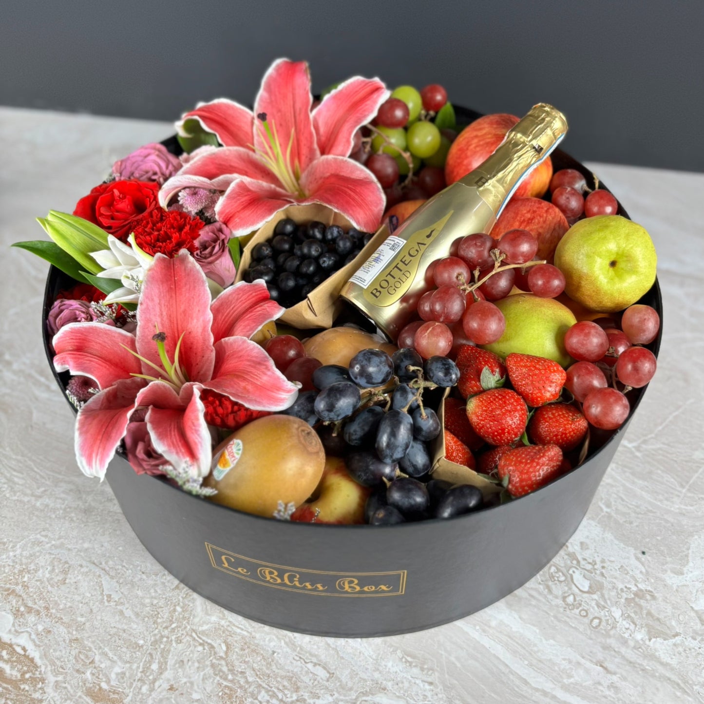 Fruit and flower arrangement in a decorative box with a bottle of champagne. Fruit hamper with flowers and a bottle of champagne on a light background. Fruit basket with flowers and a bottle of champagne on a light surface. Bottega Grand Round Fruit Box | Best Wine Fruit Hamper in Jakarta, Surabaya & Tangerang – Le Bliss Box