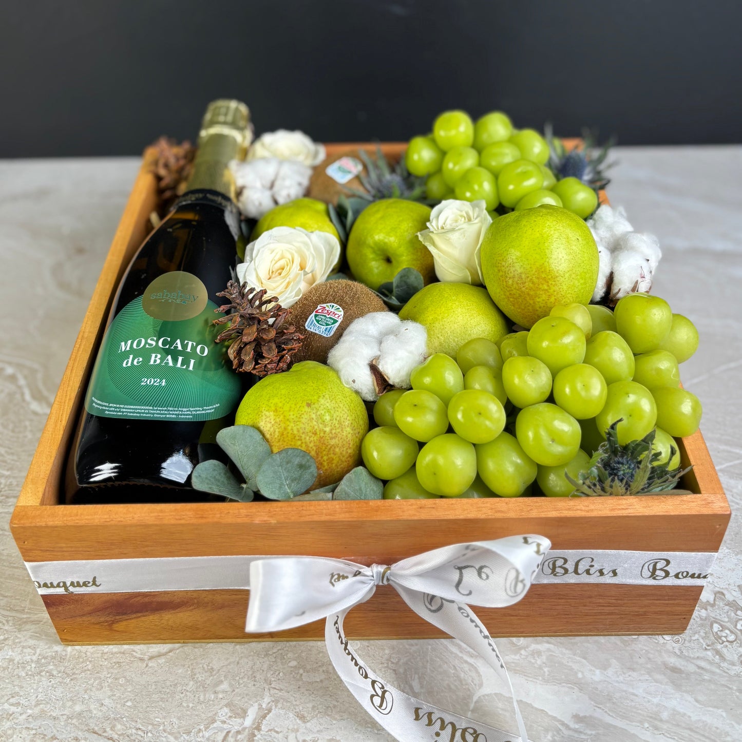 Fruit basket with a bottle of Moscato wine, green grapes, and decorative elements on a gray surface. Gift basket with Moscato de Bali wine, fruits, and flowers on a light surface. wine hamper jakarta, wine hamper surabaya by le bliss box