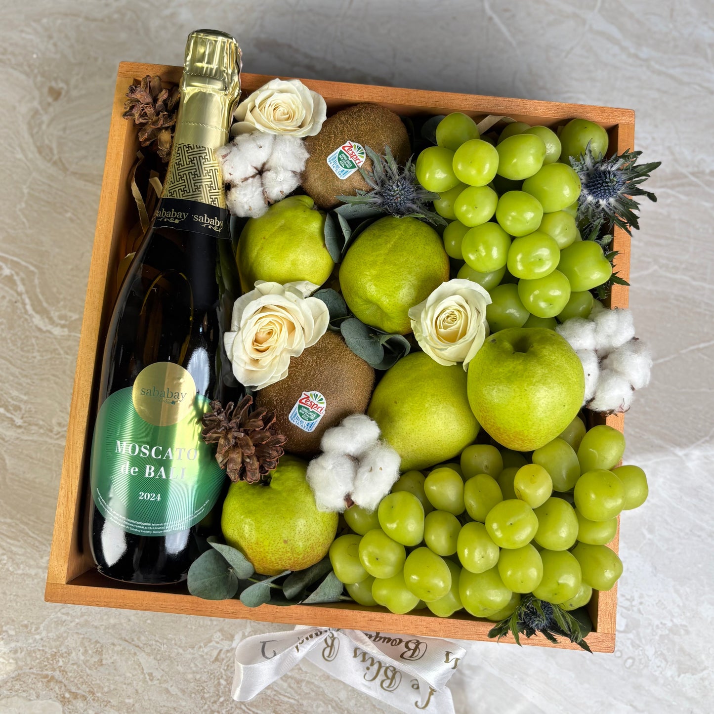 Fruit basket with a bottle of champagne on a marble surface. Gift basket with wine, muscat grapes by Le Bliss Box. Wine Sababay Hamper Jakarta, Wine hamper Surabaya