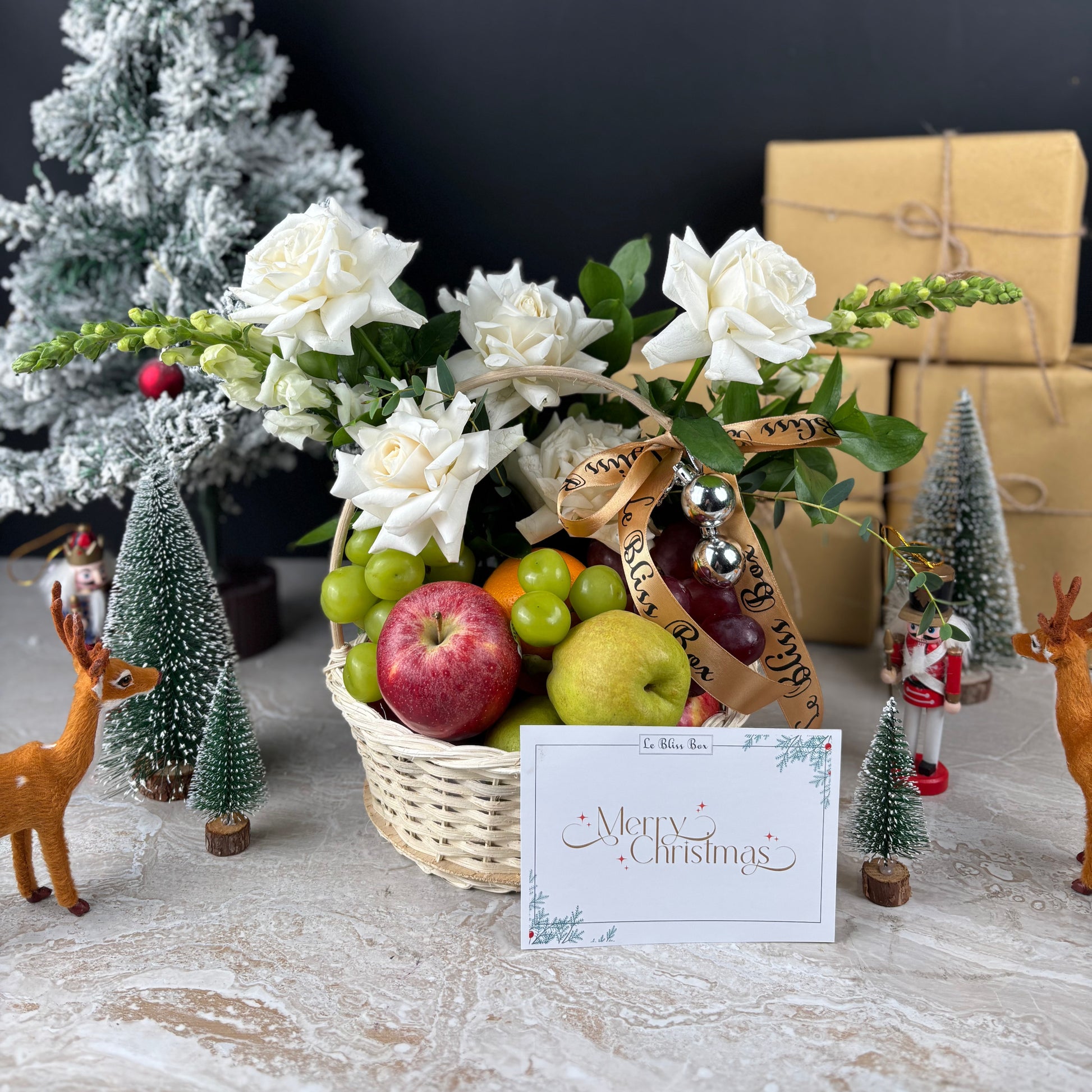 Fruit basket with flowers, Christmas decorations, and a 'Merry Christmas' card on a table. Blooming Winter Sonata Fruit Basket dari Le Bliss Box. Christmas fruit hamper mewah dengan buah premium & dekorasi festive. Pengiriman Surabaya, Jakarta & Cirebon.