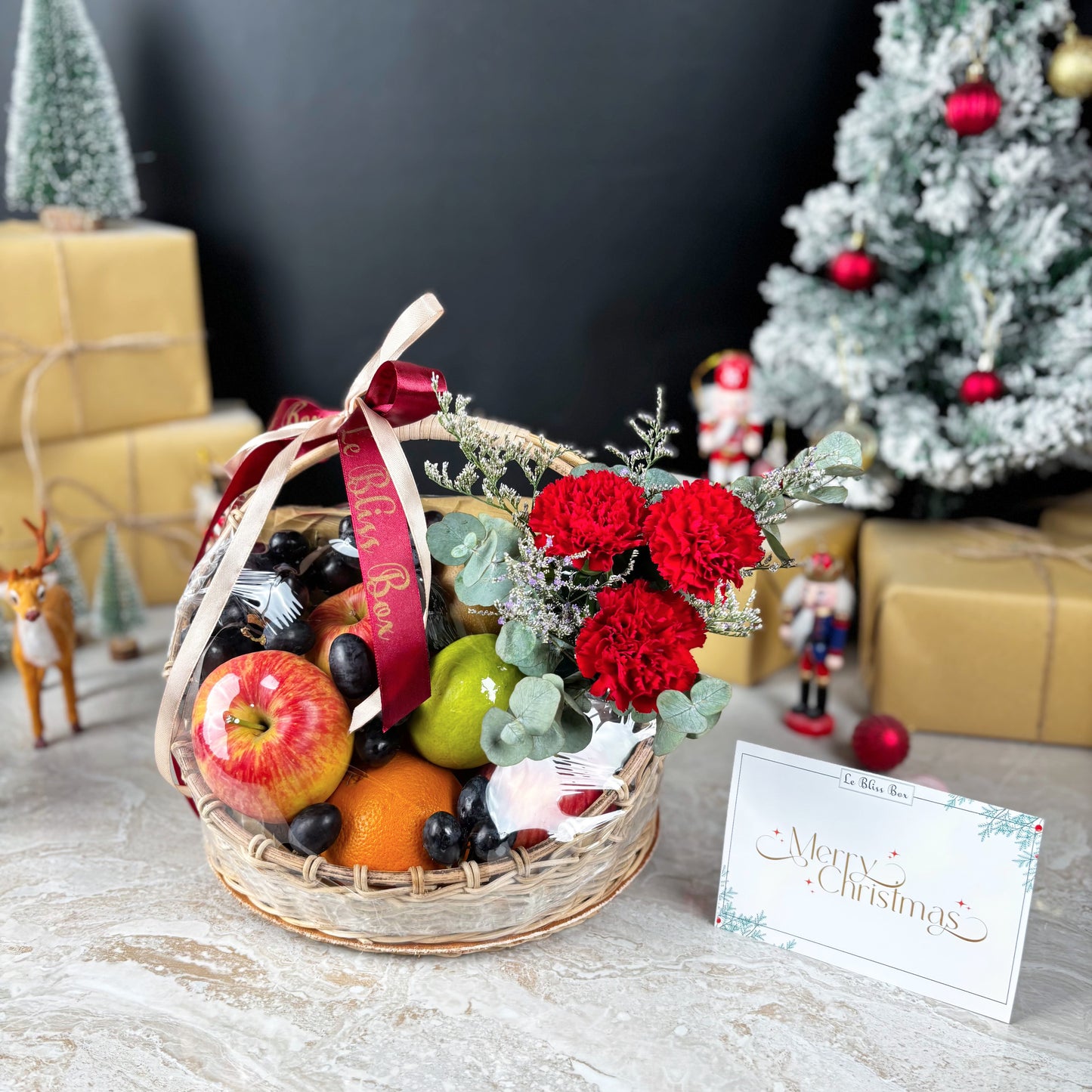 Fruit basket with flowers and a 'Merry Christmas' card in front of a decorated Christmas tree and presents. Fruit basket with flowers and a red ribbon on a festive background. Christmas Blossom Fruit Parcel | Best Christmas Hamper Jakarta, Tangerang, Cirebon & Surabaya – Le Bliss Box