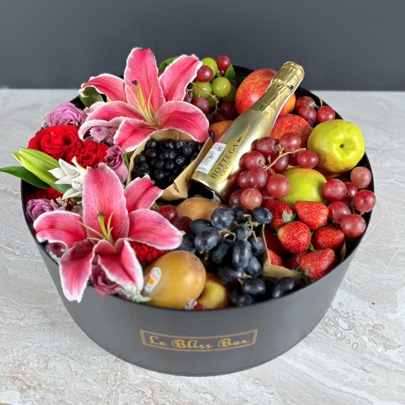Fruit basket with flowers and a bottle of champagne on a light surface. Bottega Grand Round Fruit Box | Best Wine Fruit Hamper in Jakarta, Surabaya & Tangerang – Le Bliss Box