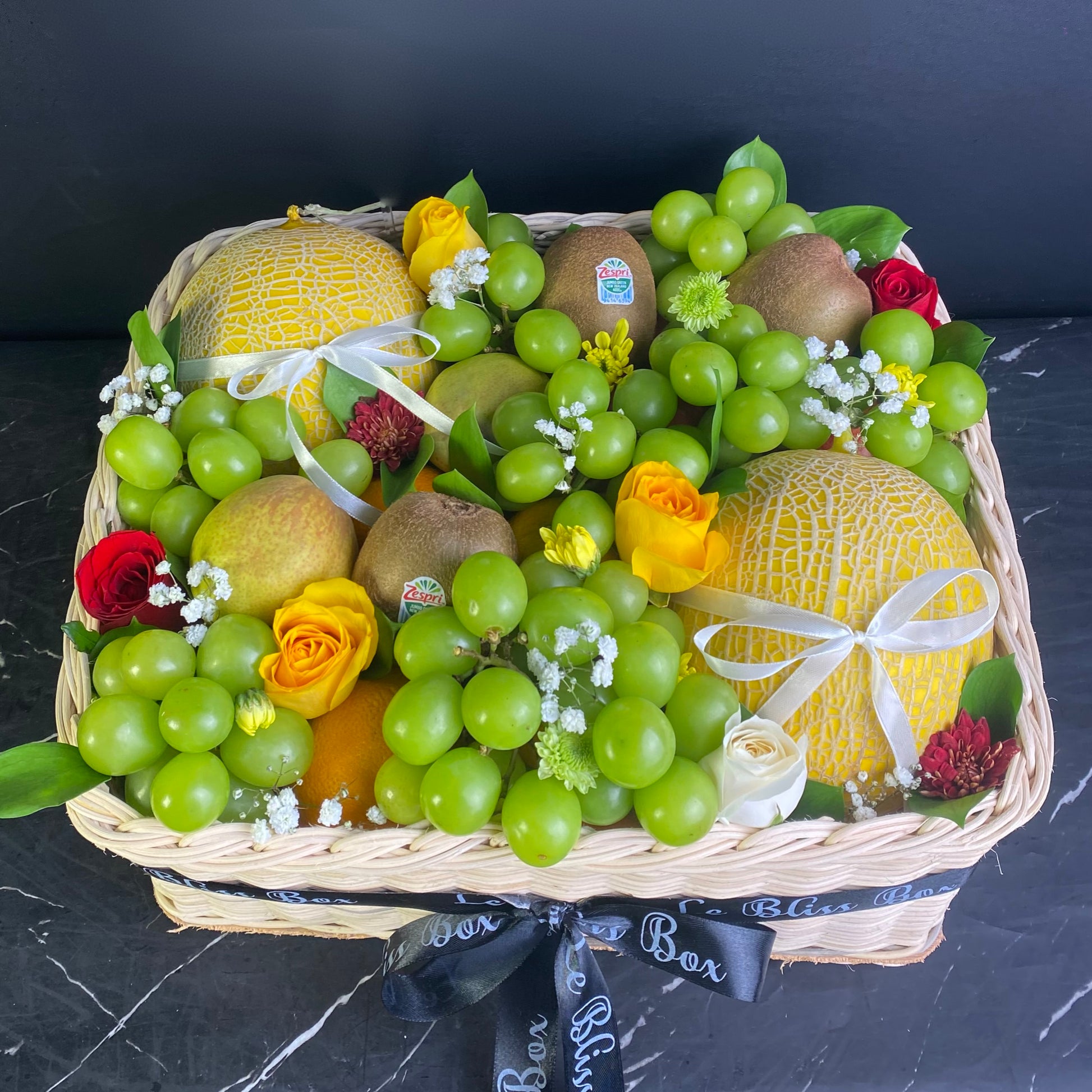 Fruit basket with grapes, melons, and flowers on a dark surface. Parcel buah melon kiwi anggur, parcel buah premium jakarta, tangerang, depok, bekasi, surabaya, gresik, cirebon