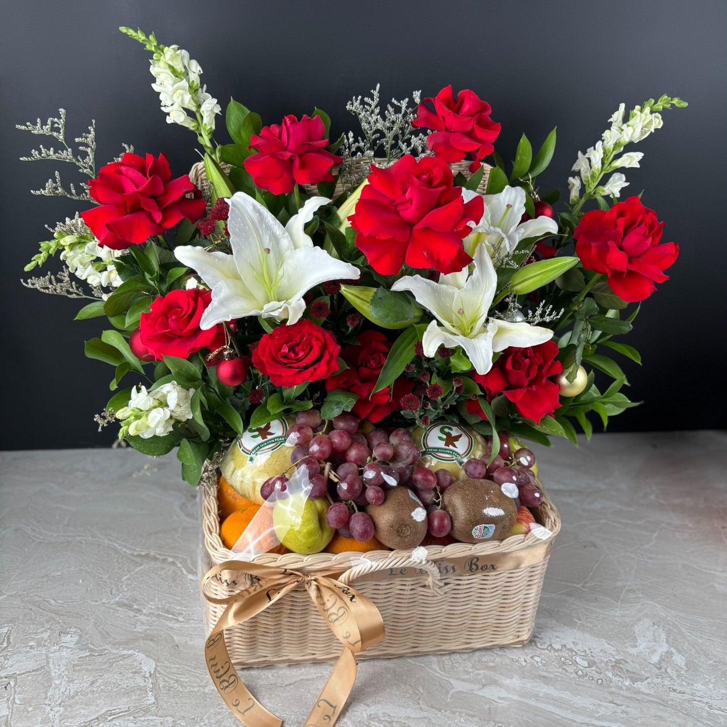 Fruit hamper of red and white flowers in a basket with fruits on a gray surface. Celebrate Christmas with Le Bliss Box’s Festive Grand Fruit Parcel — a luxurious fruit hamper for Jakarta, Surabaya & Tangerang delivery.