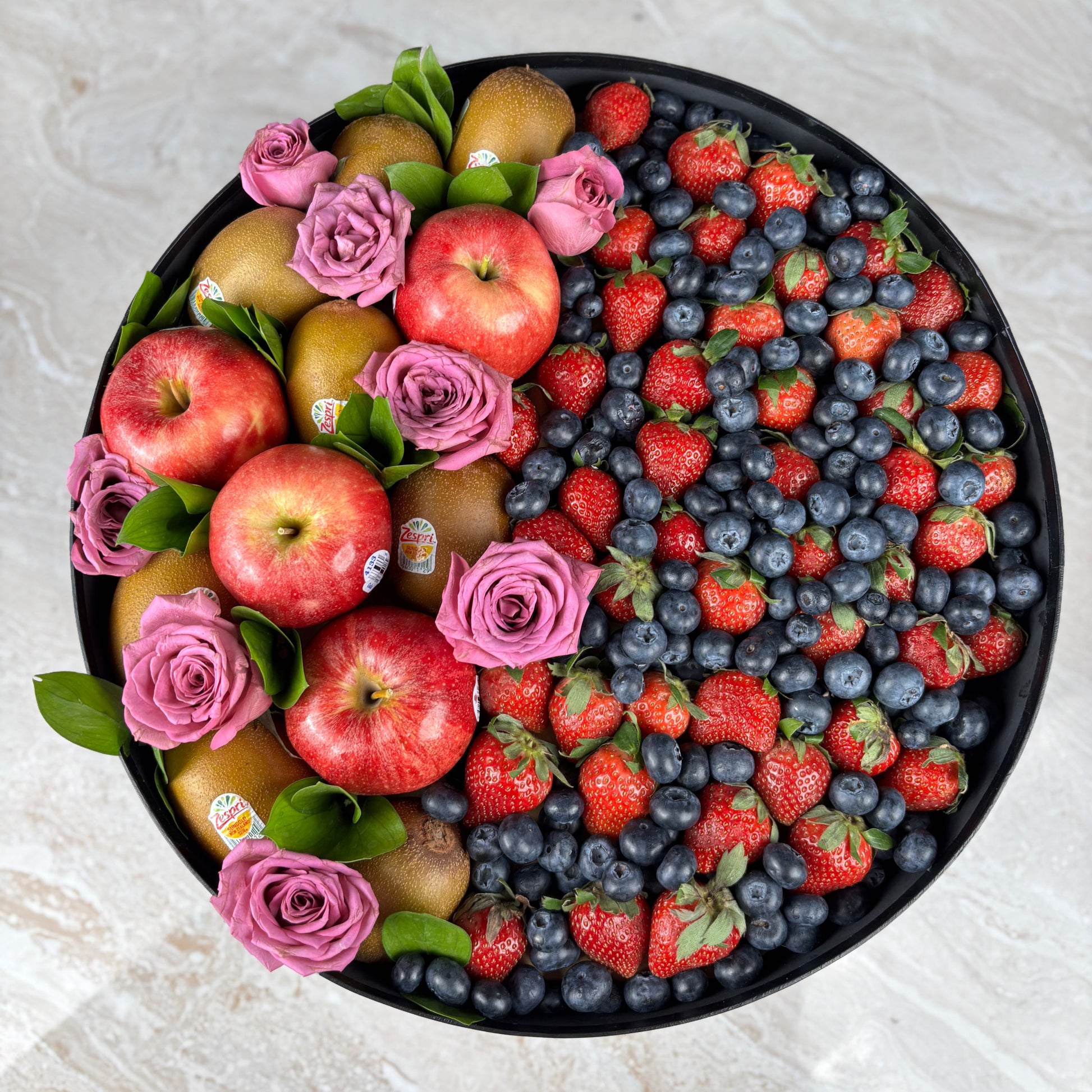 Fruit platter with apples, strawberries, blueberries, and pink roses on a light background. parcel buah terbaik surabaya, fruit hamper jakarta, kado hari ibu surabaya