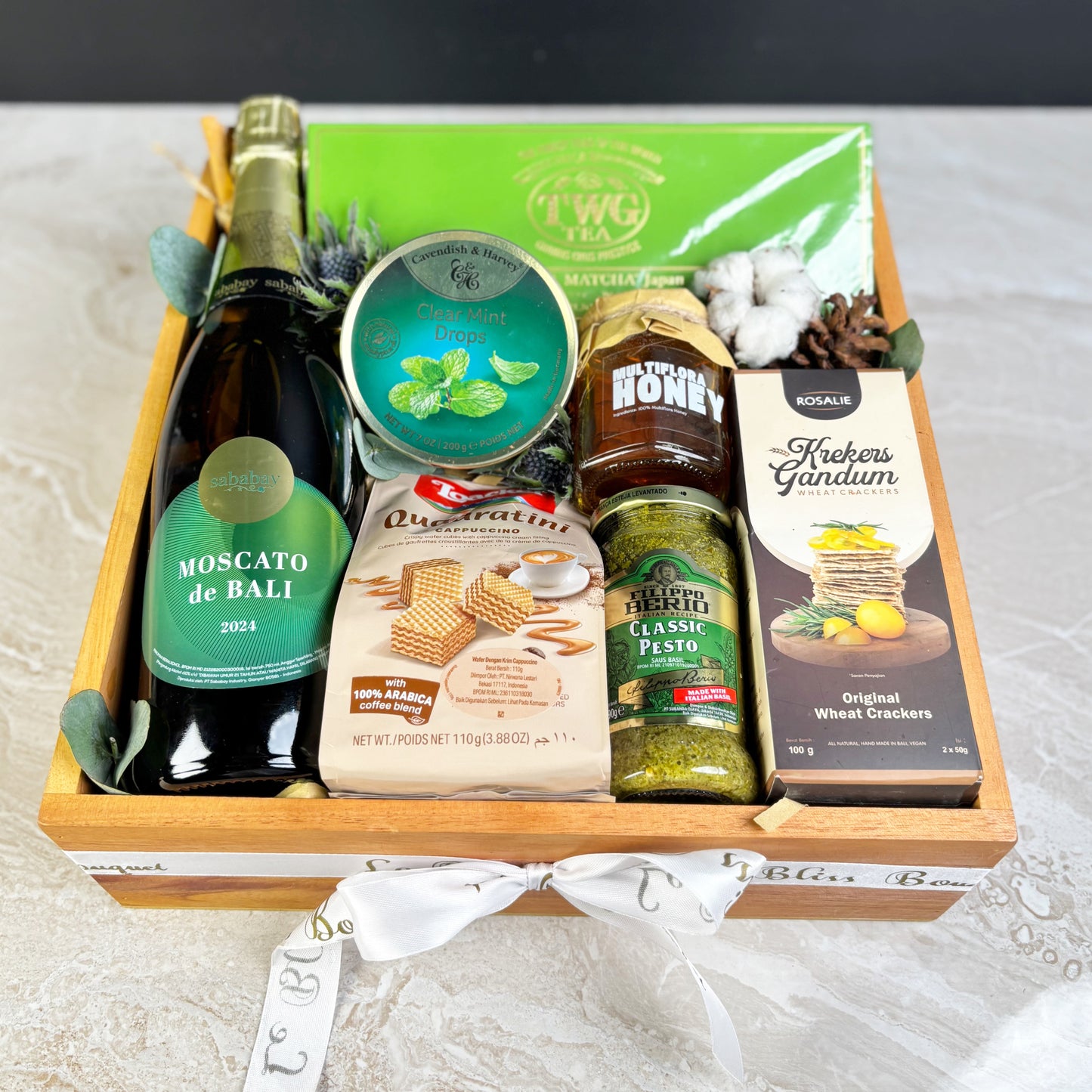 Gift basket with Moscato de Bali wine, cookies, honey, pesto, and crackers on a white surface. gourmet hamper surabaya, hamper jakarta, hamper tangerang