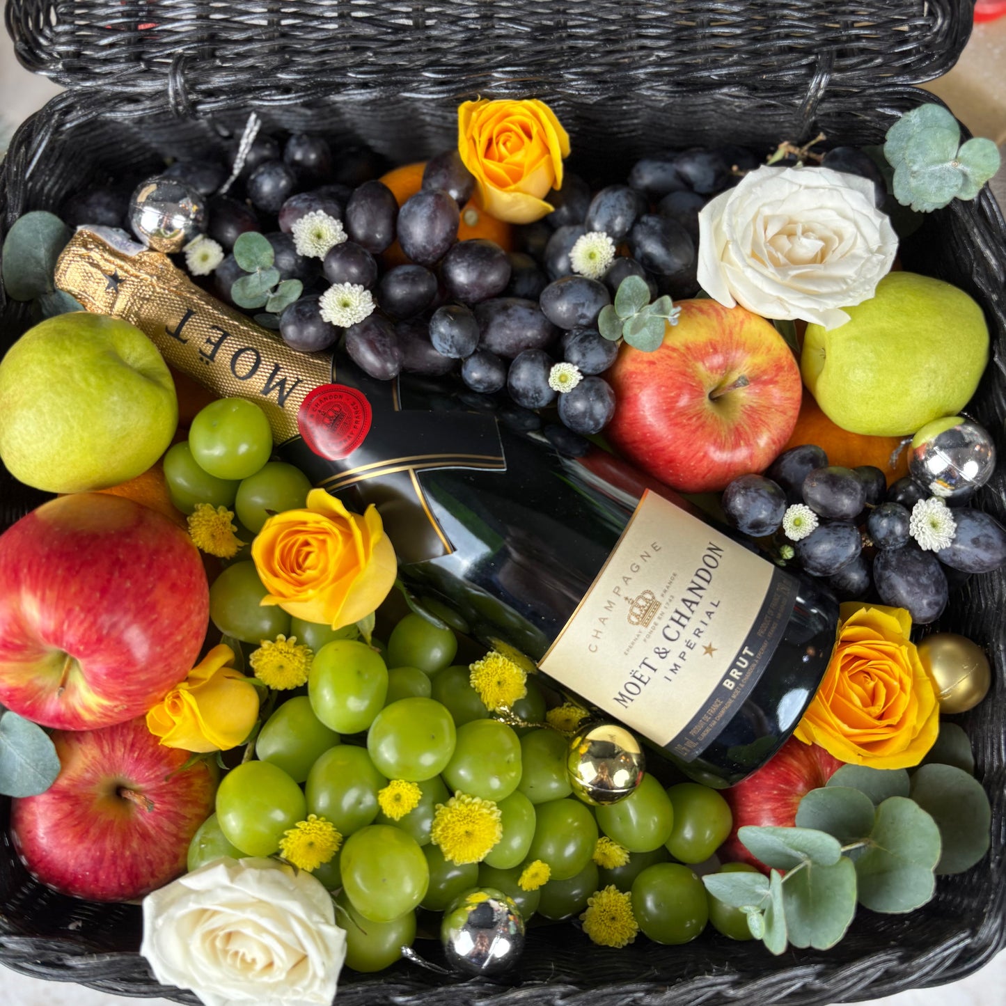 Gift basket with fruits, flowers, and a bottle of Moet & Chandon champagne. Fruit basket with moet champagne bottle, apples, grapes, and flowers on a textured surface. Midnight Moët Champagne Hamper | Best Christmas Hamper in Jakarta, Surabaya & Tangerang – Le Bliss Box