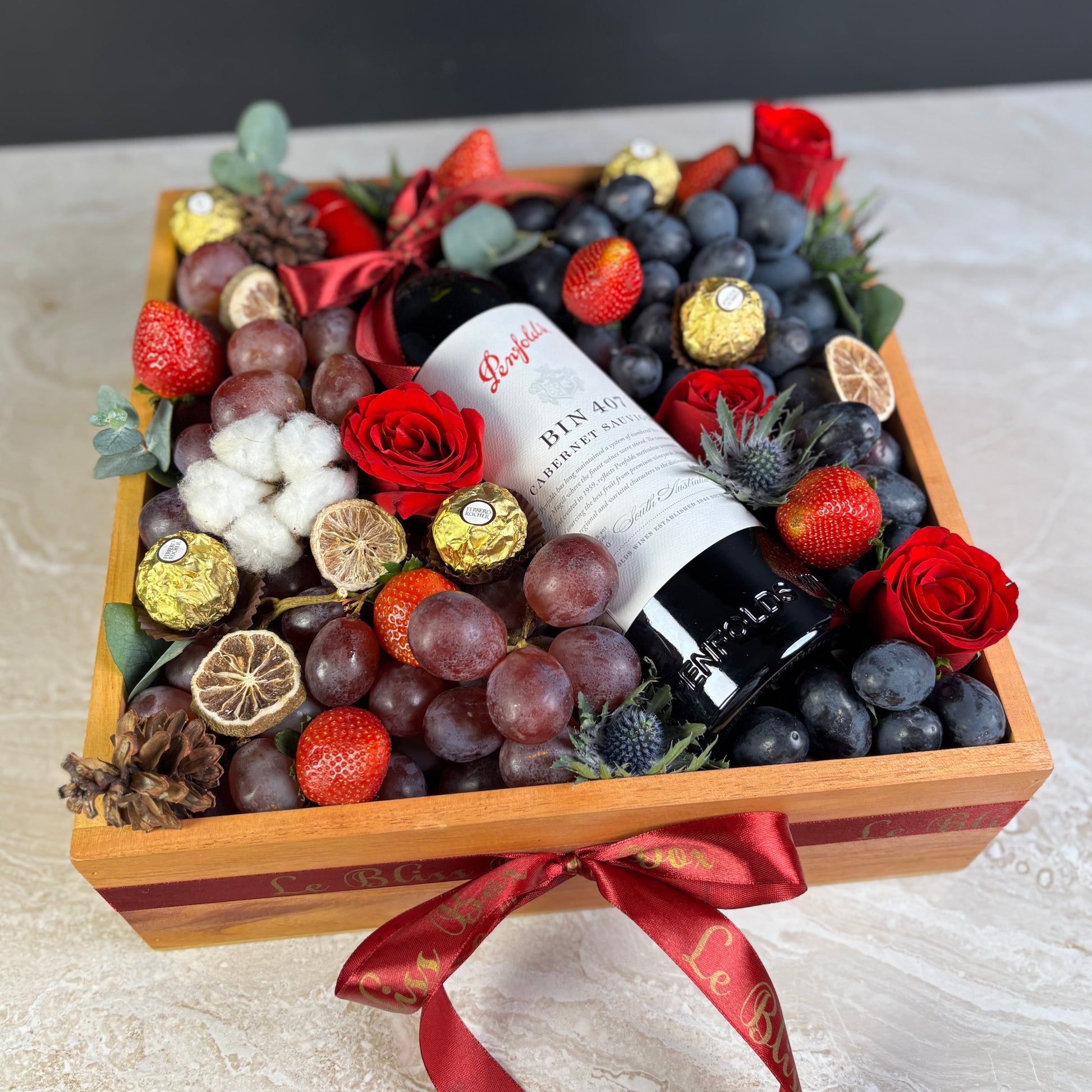 Gift basket with fruits, flowers, and a bottle of wine on a textured surface. Penfolds Wine Hamper Jakarta, Surabaya. With Fresh autumn grapes, red globe grapes and strawberries and chocolate. Wine Hamper Jakarta
