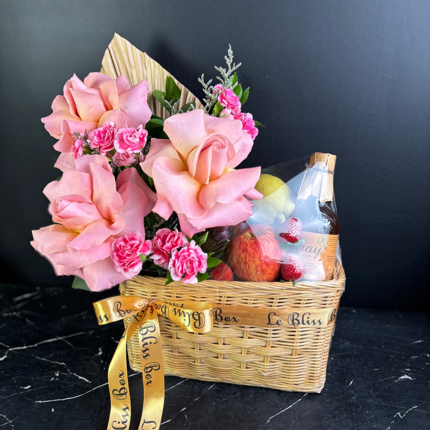 Gift basket with pink flowers, fruits, and a bottle on a dark surface. parcel buah surabaya dengan madu