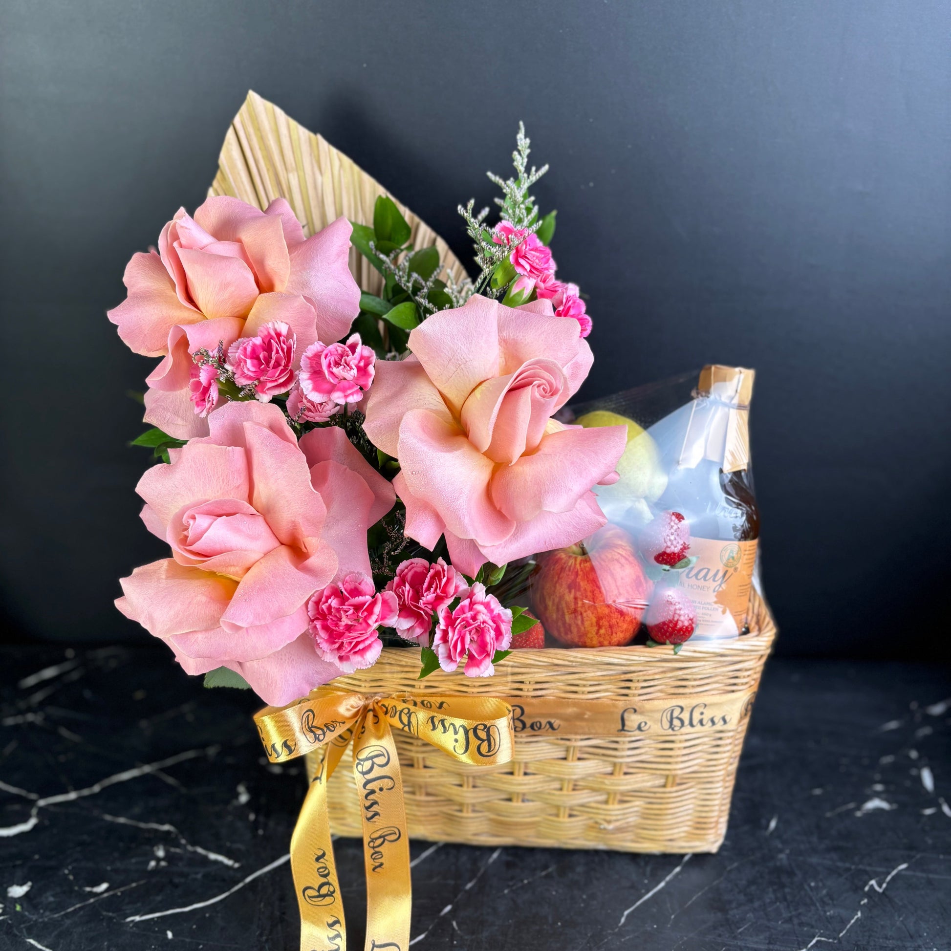 Gift basket with pink flowers, fruits, and a bottle on a dark surface. parcel buah surabaya dengan madu, fruit hamper surabaya with honey. parcel buah madu jakarta