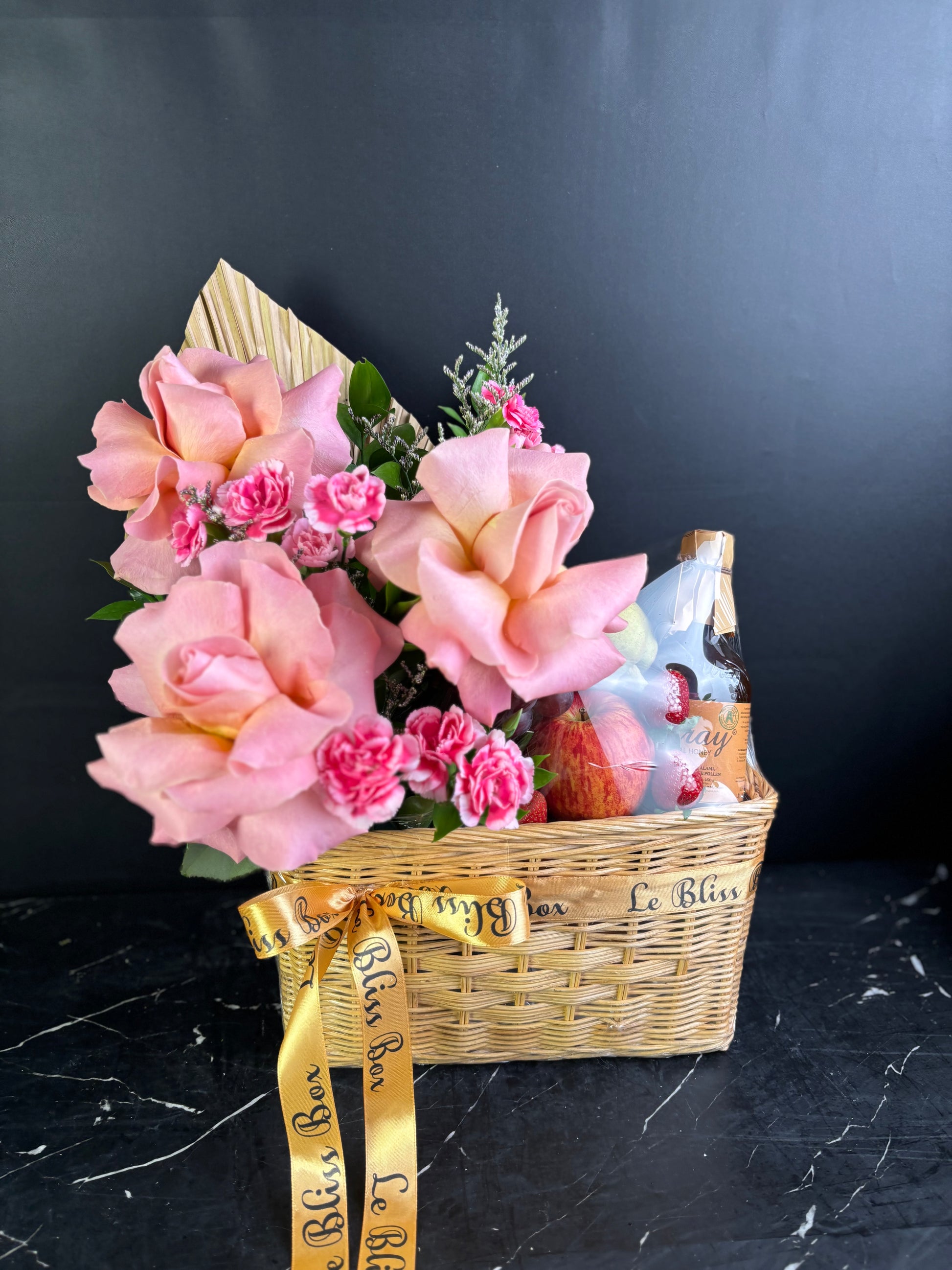 Gift basket with pink flowers, fruits, and a bottle on a dark surface. parcel buah surabaya dengan madu, fruit hamper surabaya with honey