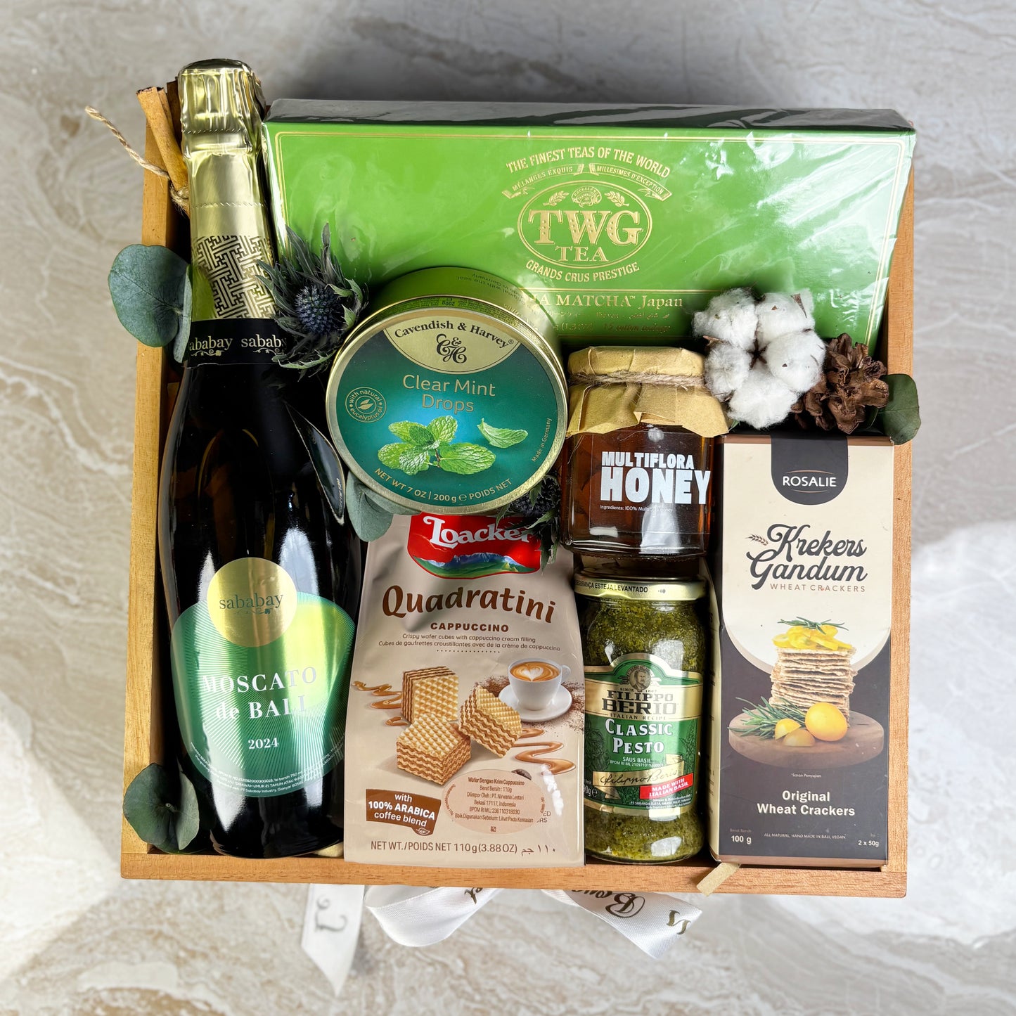 Gift basket with various items including a bottle of wine, tea, honey, and snacks on a light background. Green Serenity Gourmet Hamper dari Le Bliss Box. Parcel premium dengan pilihan gourmet mewah. Pengiriman Jakarta, Surabaya, Tangerang & Cirebon. Elegant and refined.