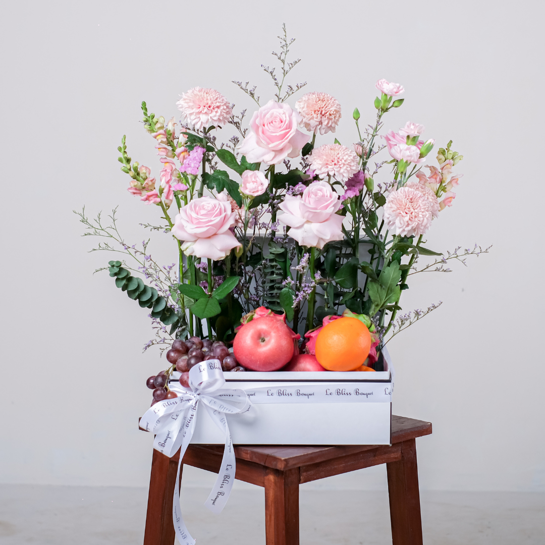 Parcel buah yang indah dari Le Bliss Bouquet, menampilkan buah segar seperti delima, anggur, dan jeruk, dipadukan dengan rangkaian bunga mawar pink, anyelir, dan bunga lainnya yang elegan, disusun rapi dalam kotak hadiah putih dengan pita.