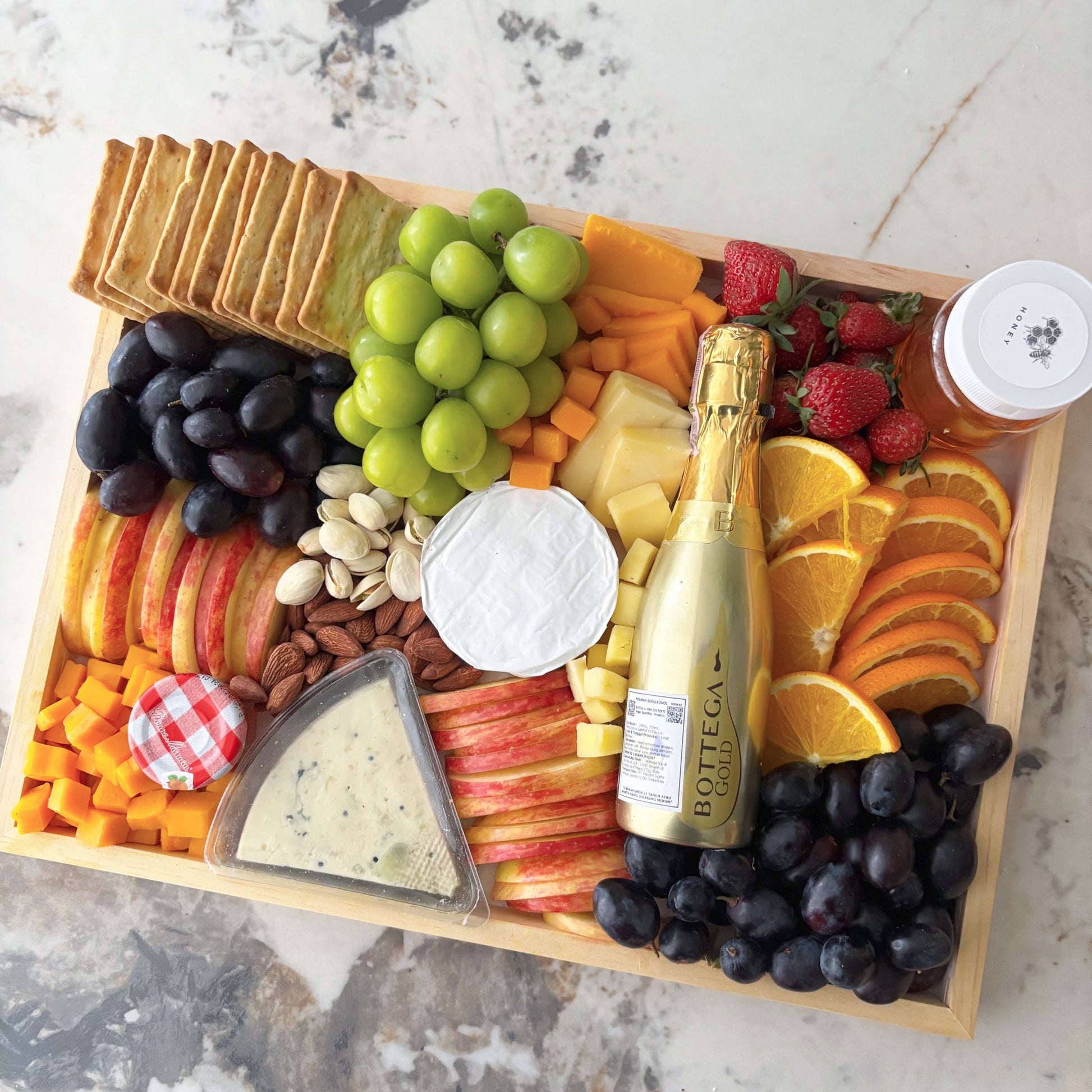 Gourmet charcuterie board with fruits, cheese, and a bottle of champagne on a marble surface. Assorted fruits, cheeses, and crackers on a wooden board with a bottle of champagne. Bottega Wine Cheese Platter | Best Wine & Cheese Platter in Jakarta & Surabaya – Le Bliss Platter